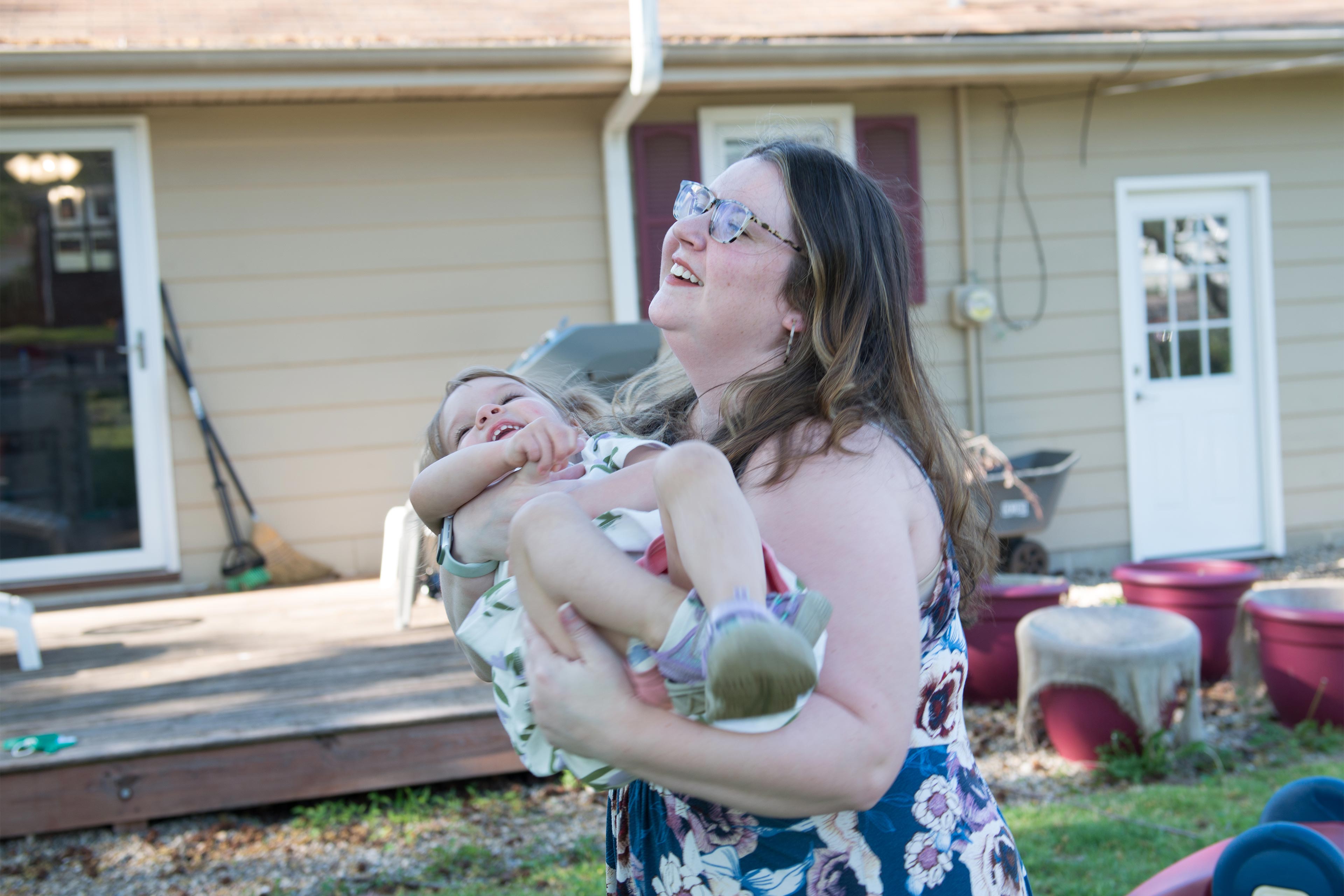 A photo of a mother holding her toddler and laughing outside.
