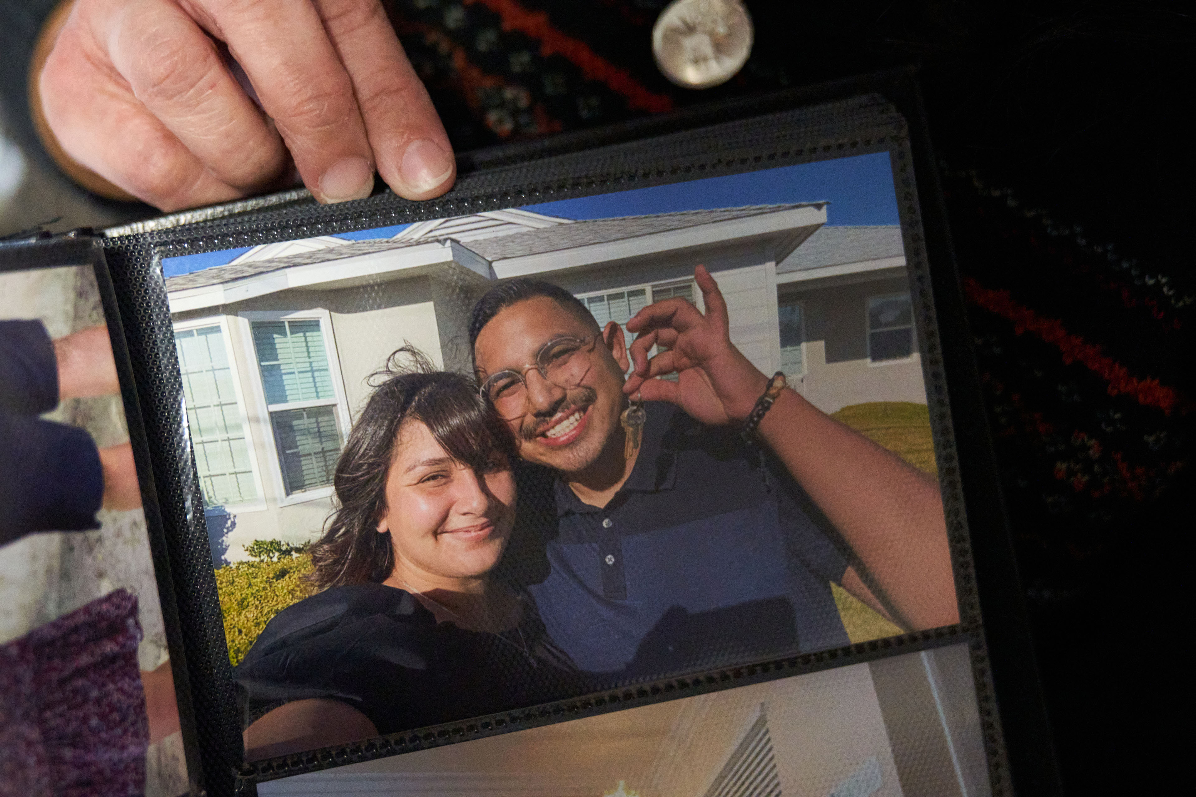 María Rivas Cruz holds up a photo in an album to the camera. It shows a photo of her and Raymond Olivares smiling in front of a house they just bought together. Olivares holds the house key up with a big smile.