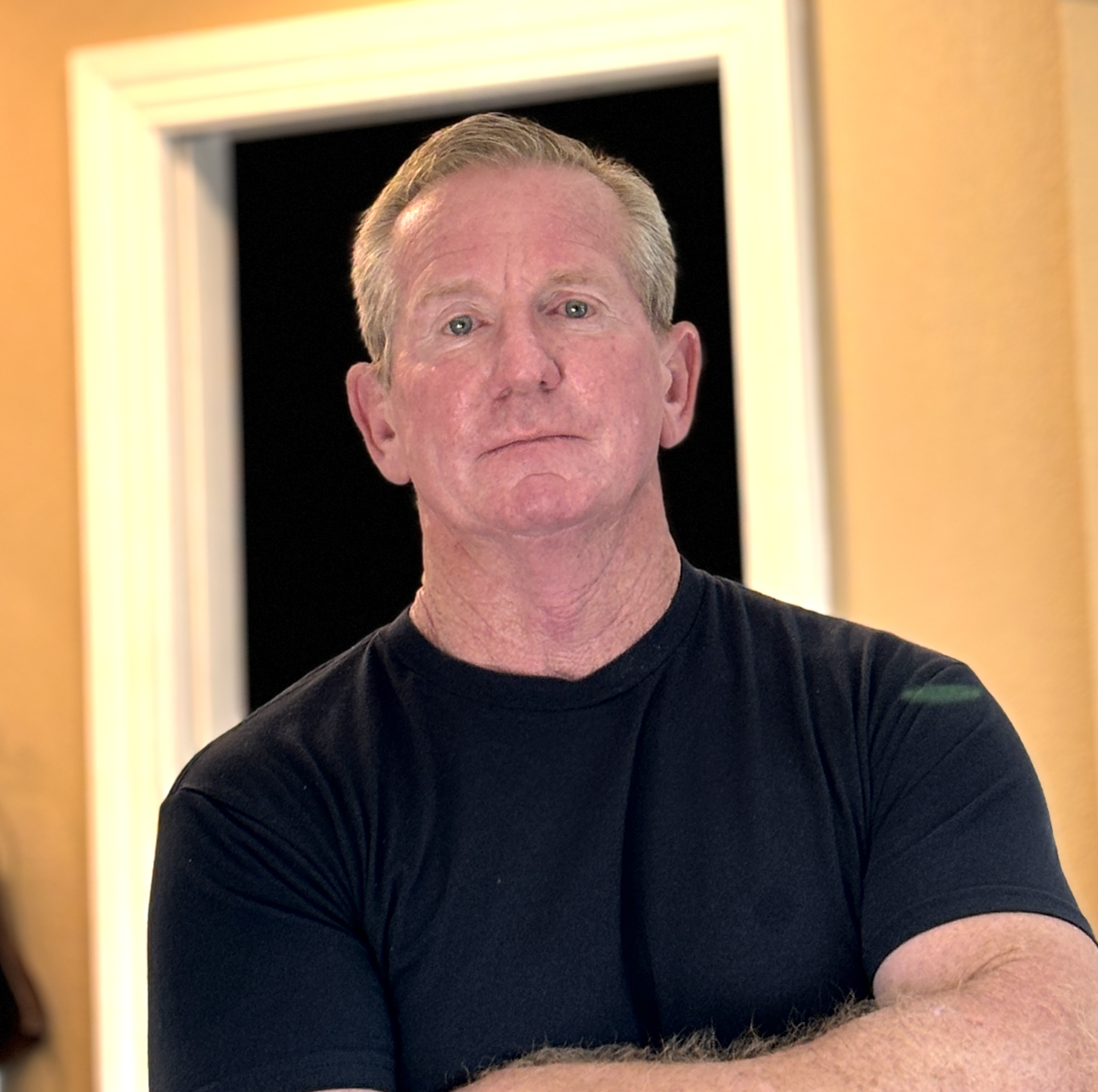 A man in a black t-shirt stands with one arm crossed in front of him and looks at the camera.