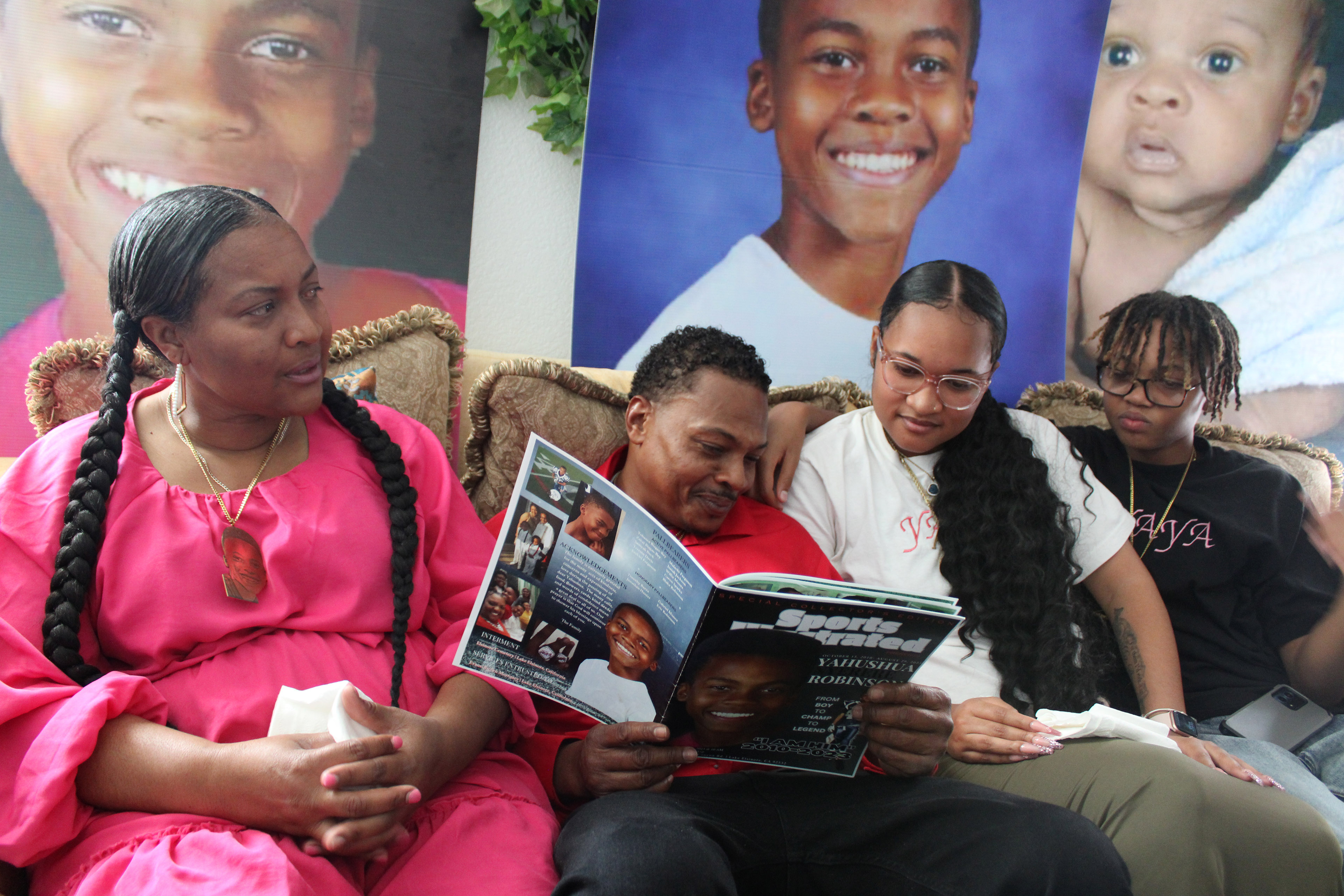 Janee and Eric Robinson sit on the couch with their two children. Together, they look at a photo album that Eric is holding. Behind them are large photographs of Yahushua Robinson at different ages.