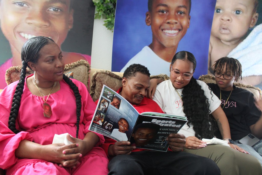 Janee and Eric Robinson sit on the couch with their two children. Together, they look at a photo album that Eric is holding. Behind them are large photographs of Yahushua Robinson at different ages.