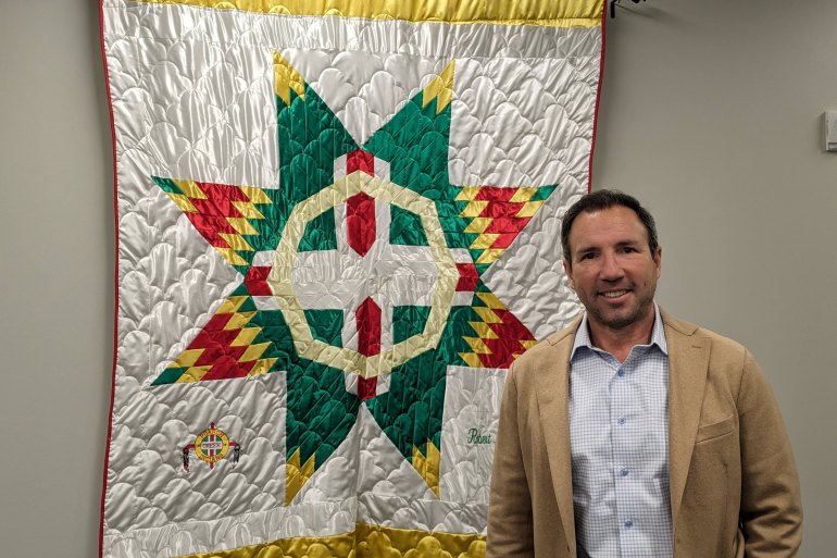 A man in a tan suit jacket smiles at the camera while standing in front of a quilted piece of white, yellow, green, and red fabric mounted to the wall behind him.