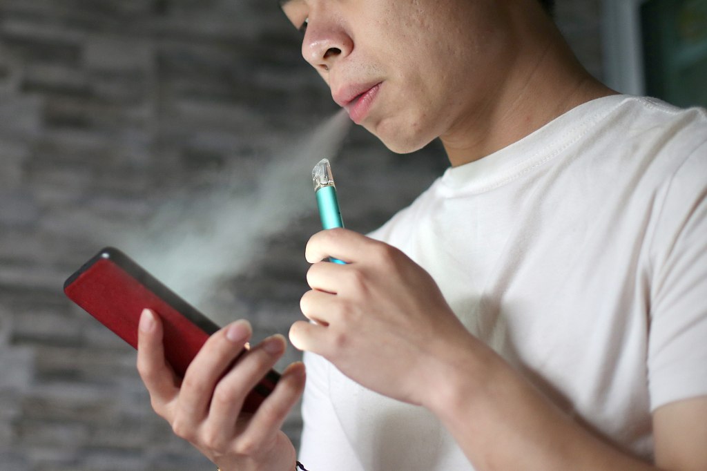 A photo of a young man vaping while looking at his phone.