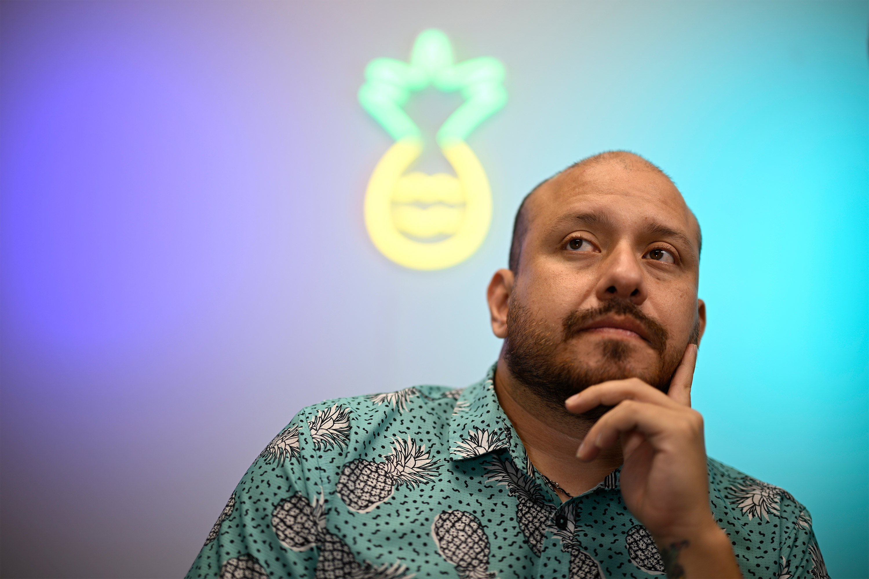 A photo of a man sitting indoors in front of wall lit with colorful lights — including a pineapple-shaped light.