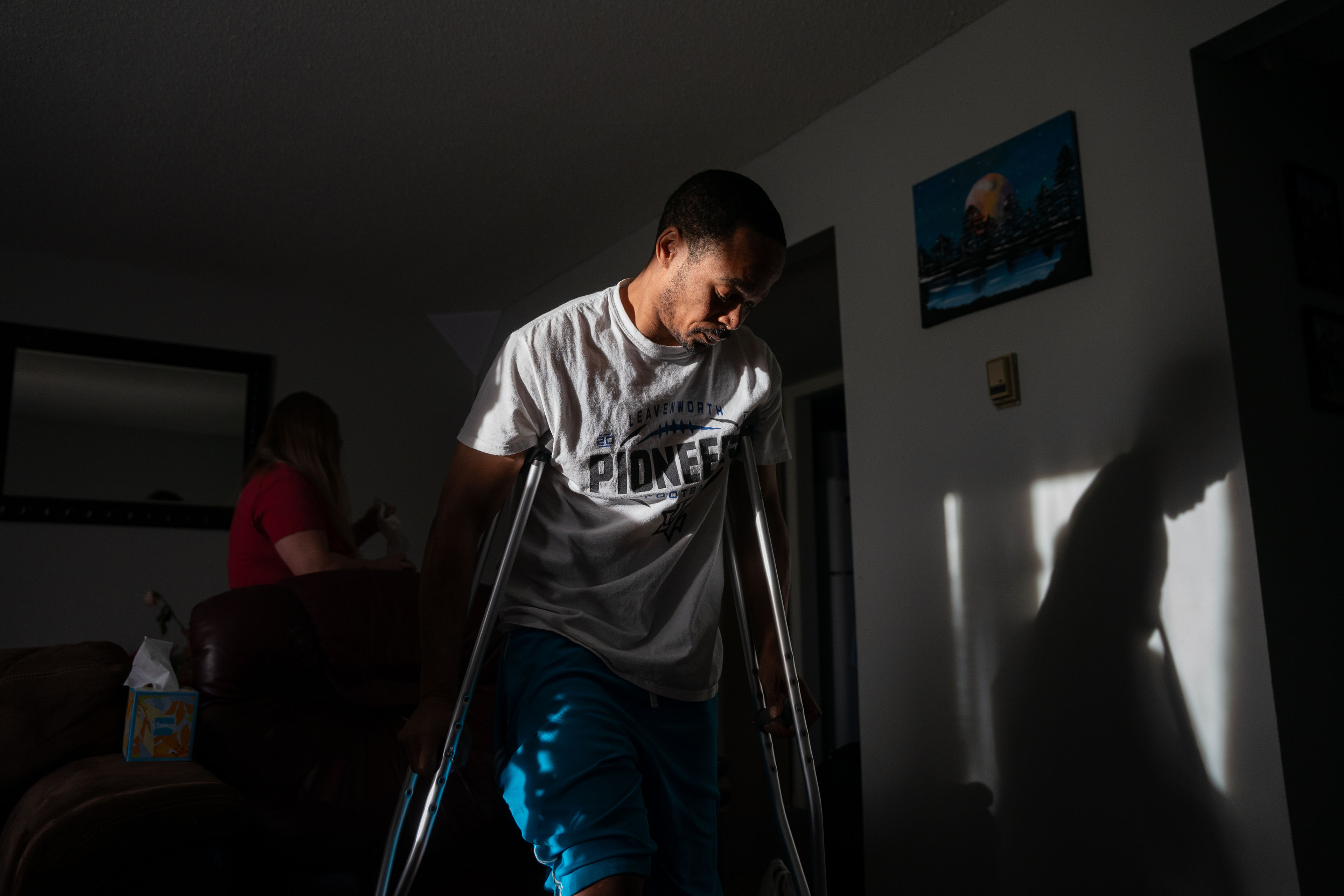 Jacob Gooch Sr. is standing on crutches in a room in his home. The room is dark, but he stands in a beam of light coming in through a window.