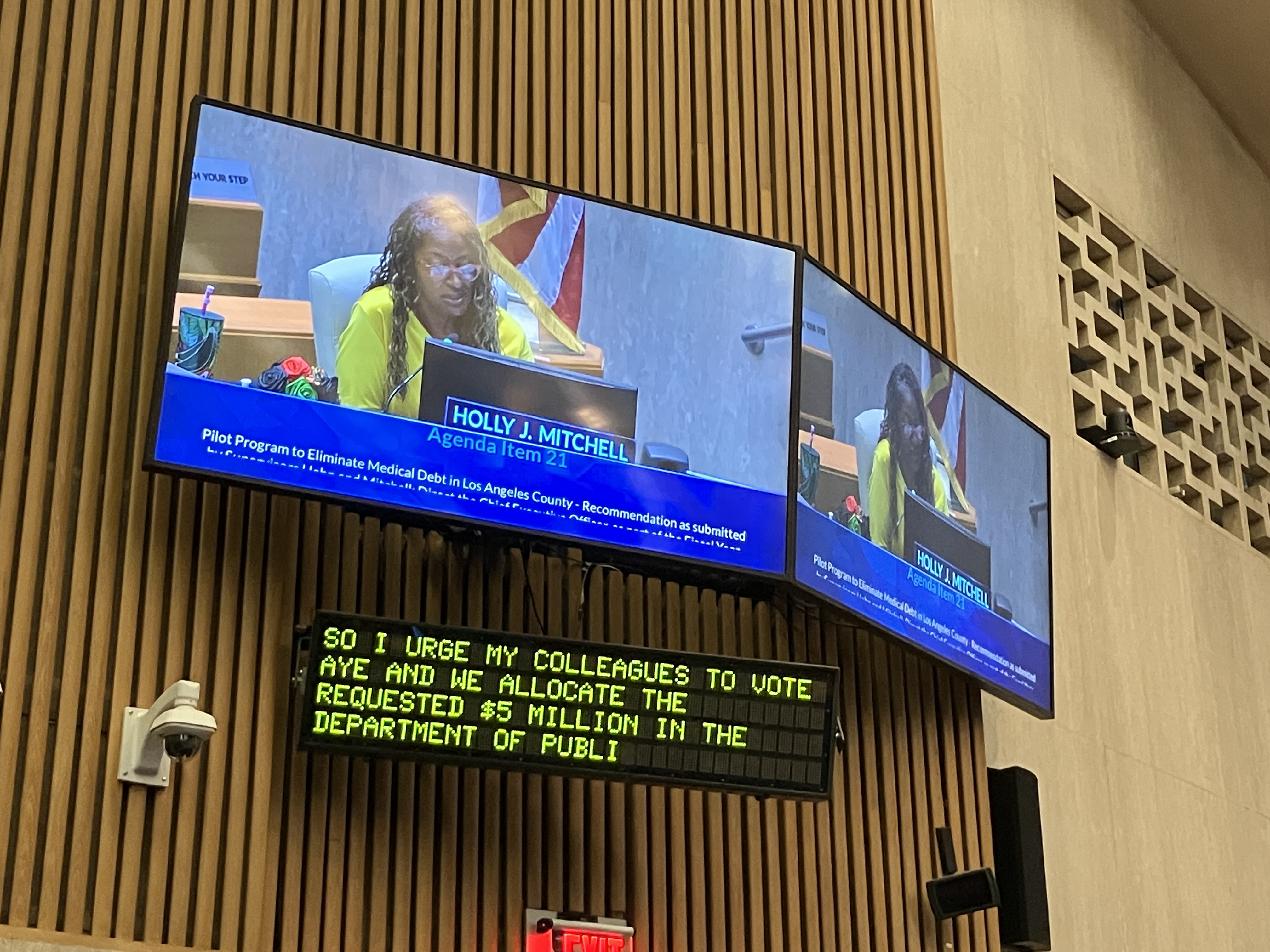 Los Angeles County Supervisor Holly Mitchell is shown on a TV screen in the court room. A screen below the TV shows in text that she is saying, "So I urge my colleages to vote aye and we allocate the requested $5 million in the Department of Publi[c]..."