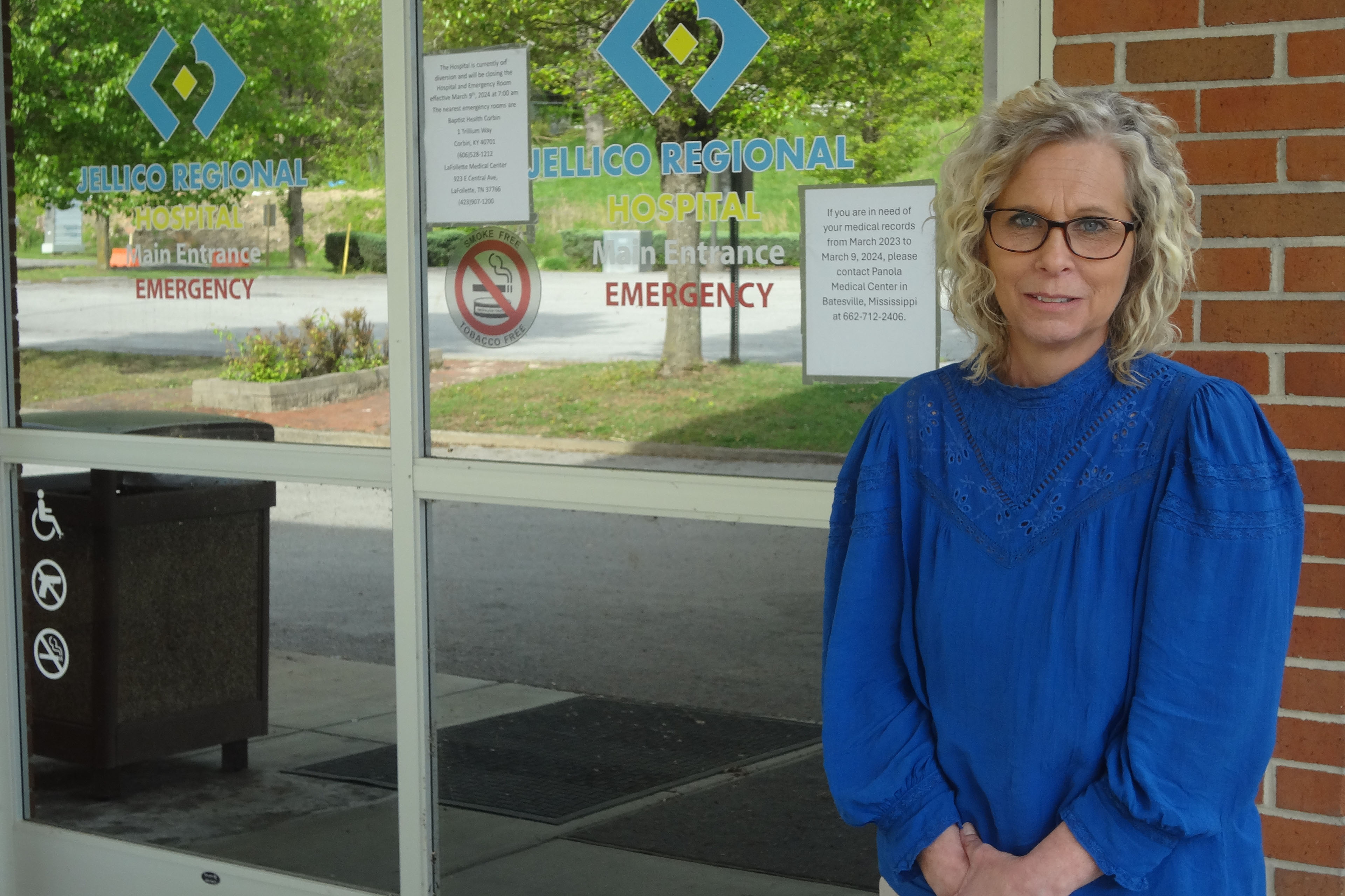 Sandy Terry, wearing a vivid blue blouse, stands beside glass doors that are the entrance to the former Jellico Medical Center.