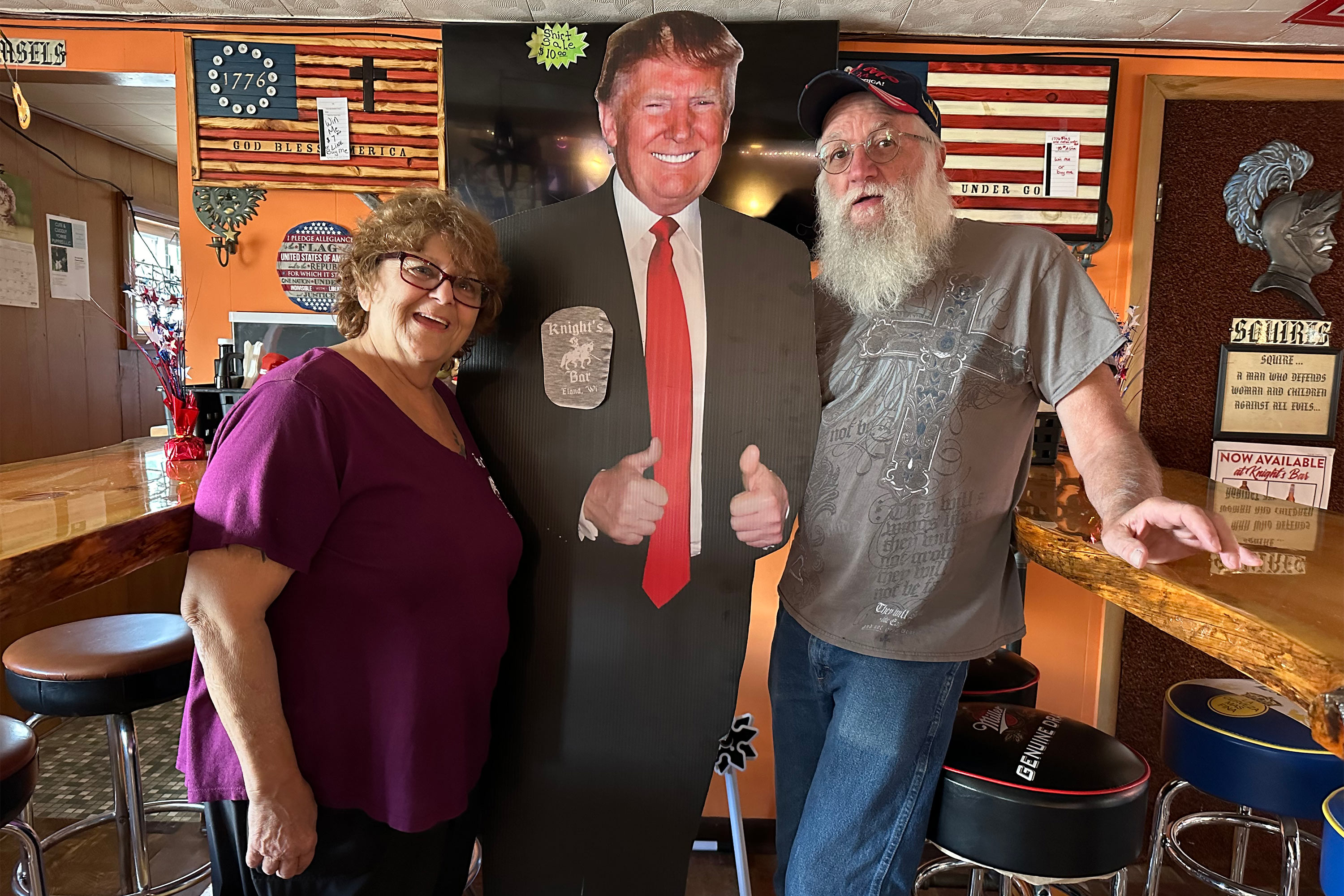 A photo of a couple posing for a couple with a life-sized cutout of former President Donald Trump indoors.