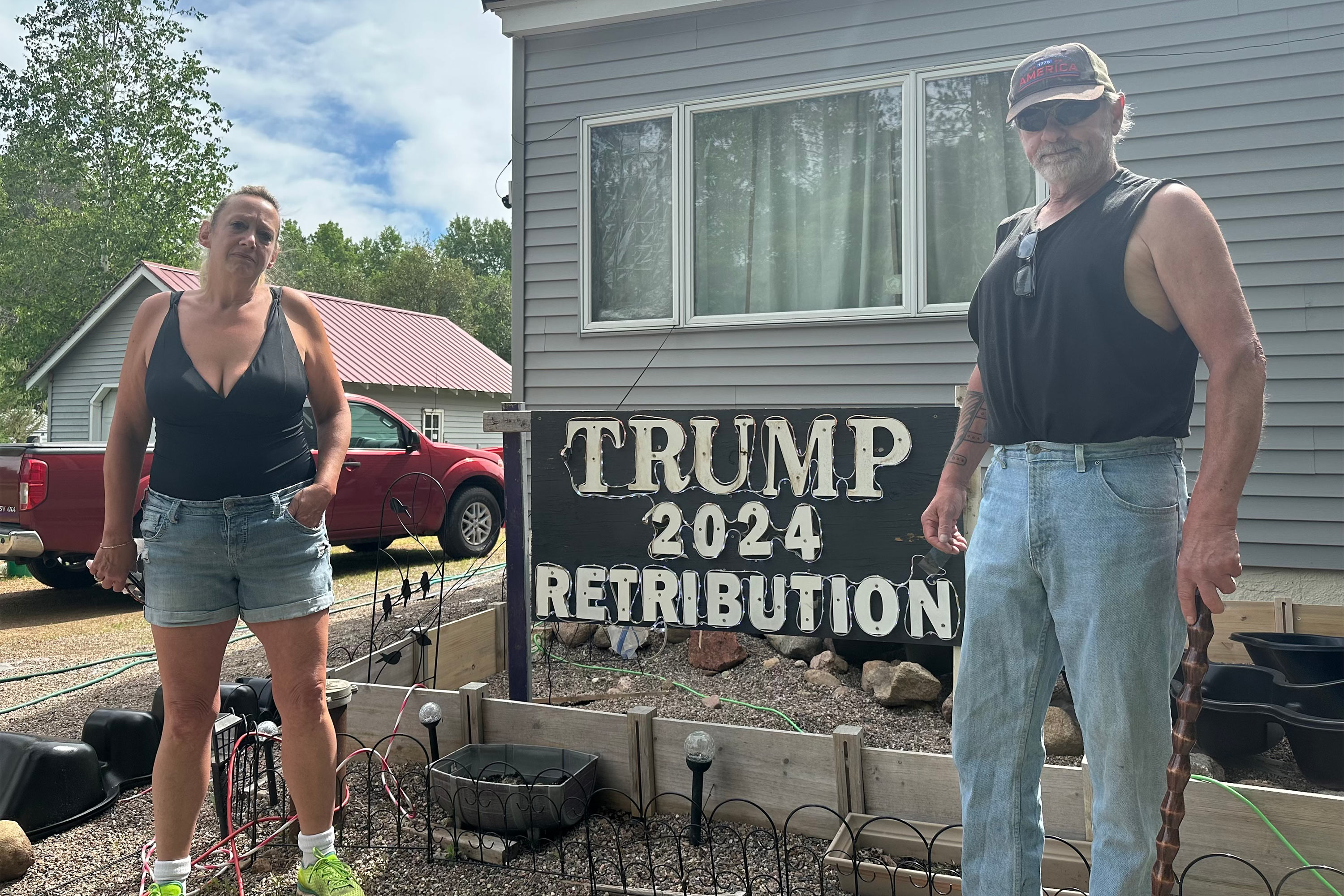 A photo of a couple standing outside. Between them is a sign that reads "Trump 2024: Retribution."
