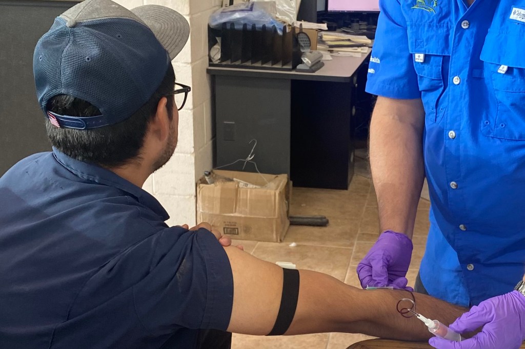 A man in a dark blue shirt and cap faces away from the camera and stretches out his right arm where another person wearing purple medical gloves draws blood.