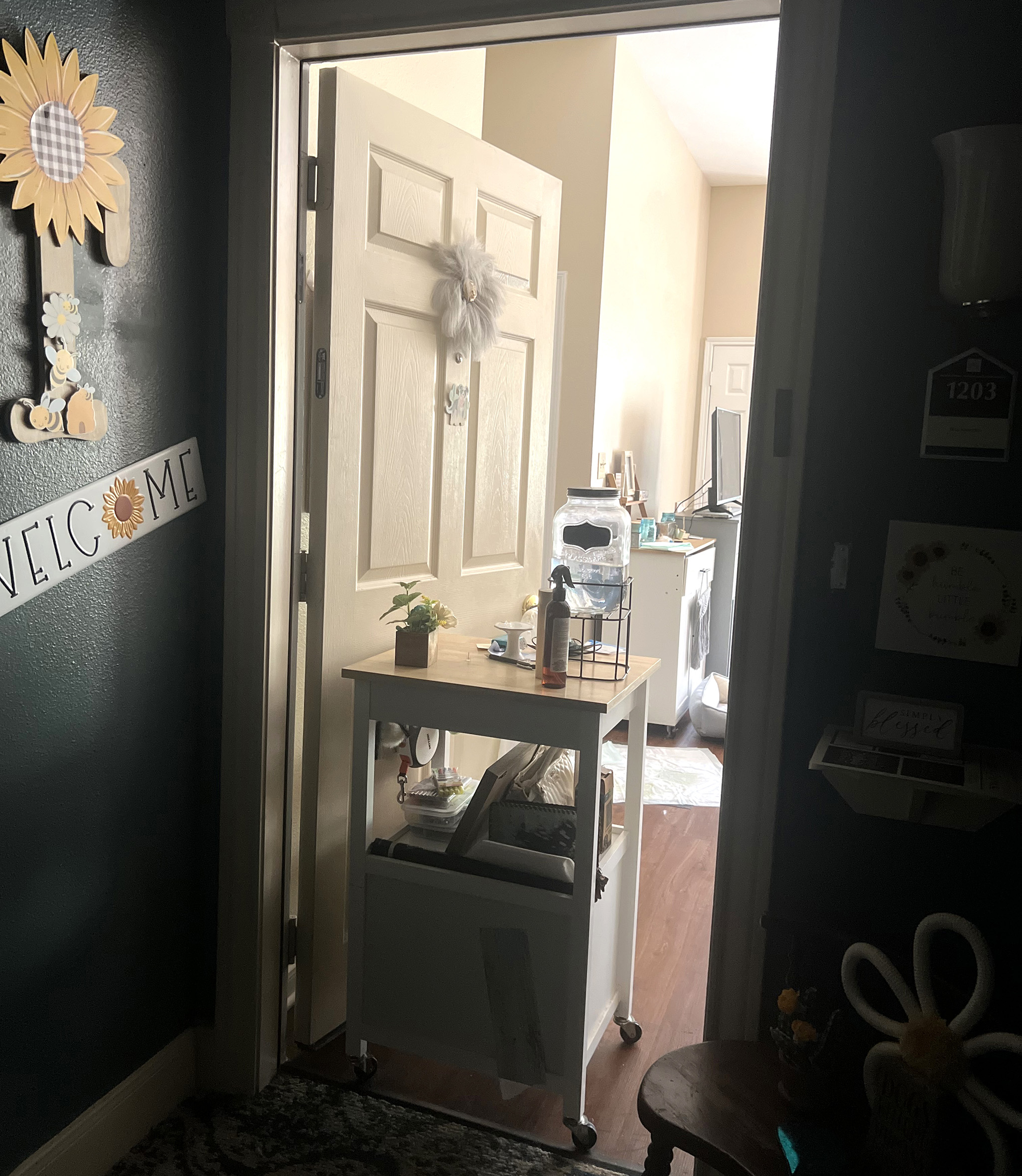 A welcome sign and sunflower hang on a hallway wall next to an open apartment door with a rolling cart holding the door open