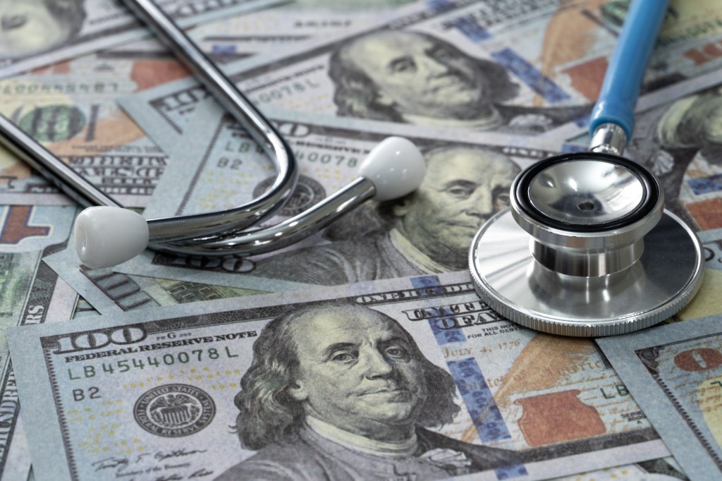 A stethoscope entwined with stacks of hundred-dollar bills on a blue background.
