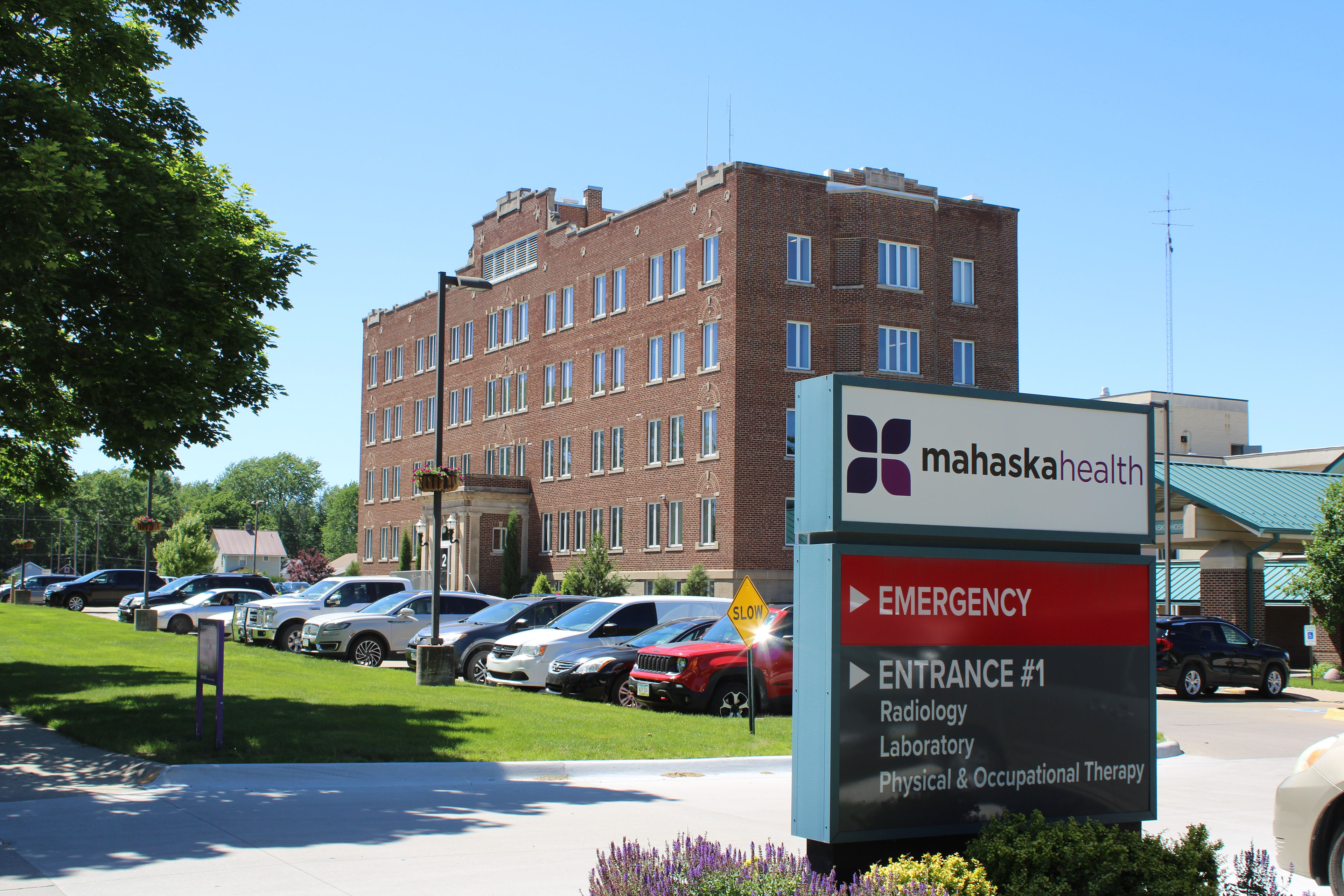 The exterior of the Mahaska Health hospital building. It is a brick building with a parking lot and bright green lawn in front. An entrance sign is stationed to the right.