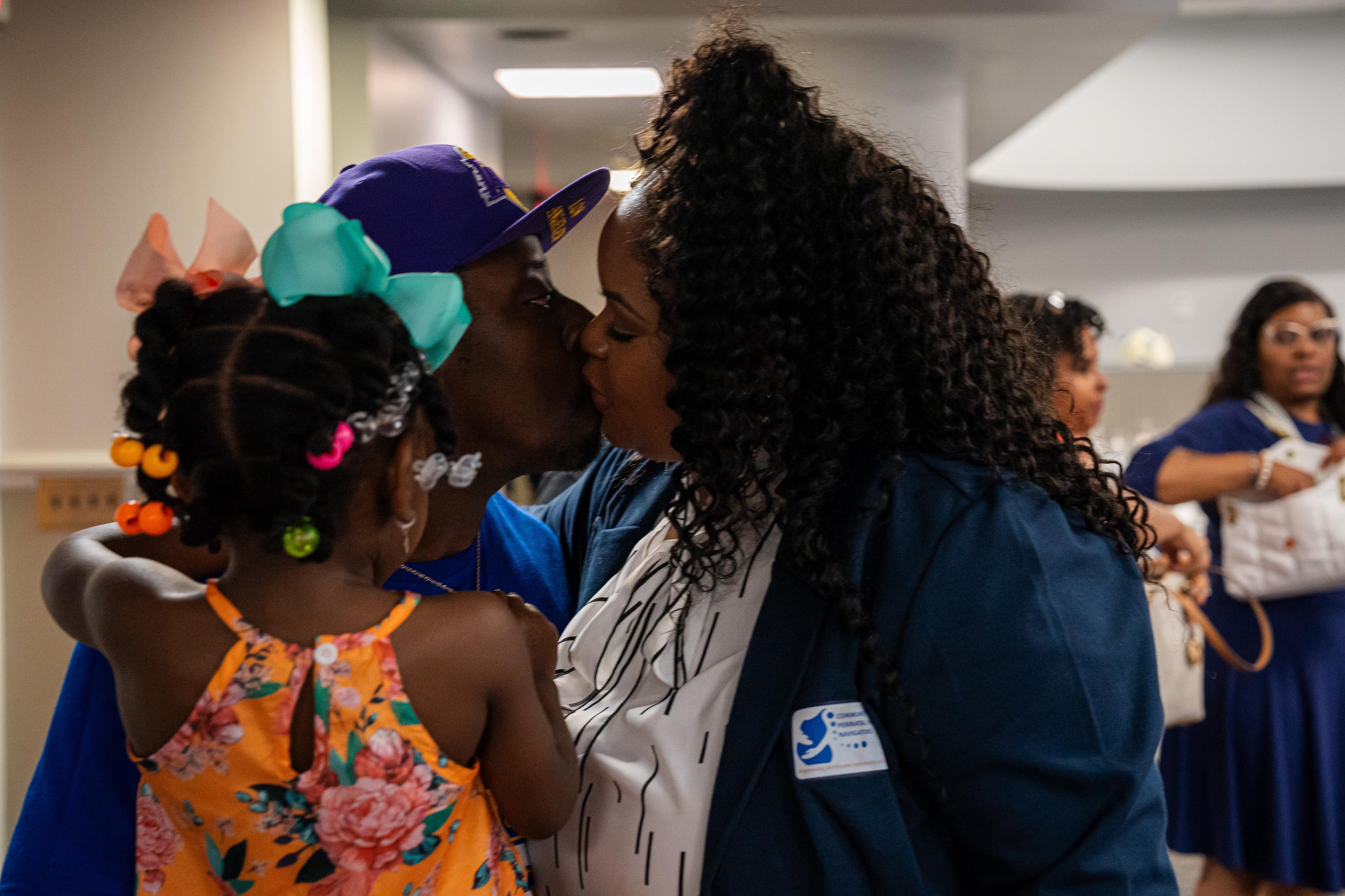 A woman wearing a blue blazer kisses a man, who is wearing in a purple baseball hat and holding a young girl in his arms