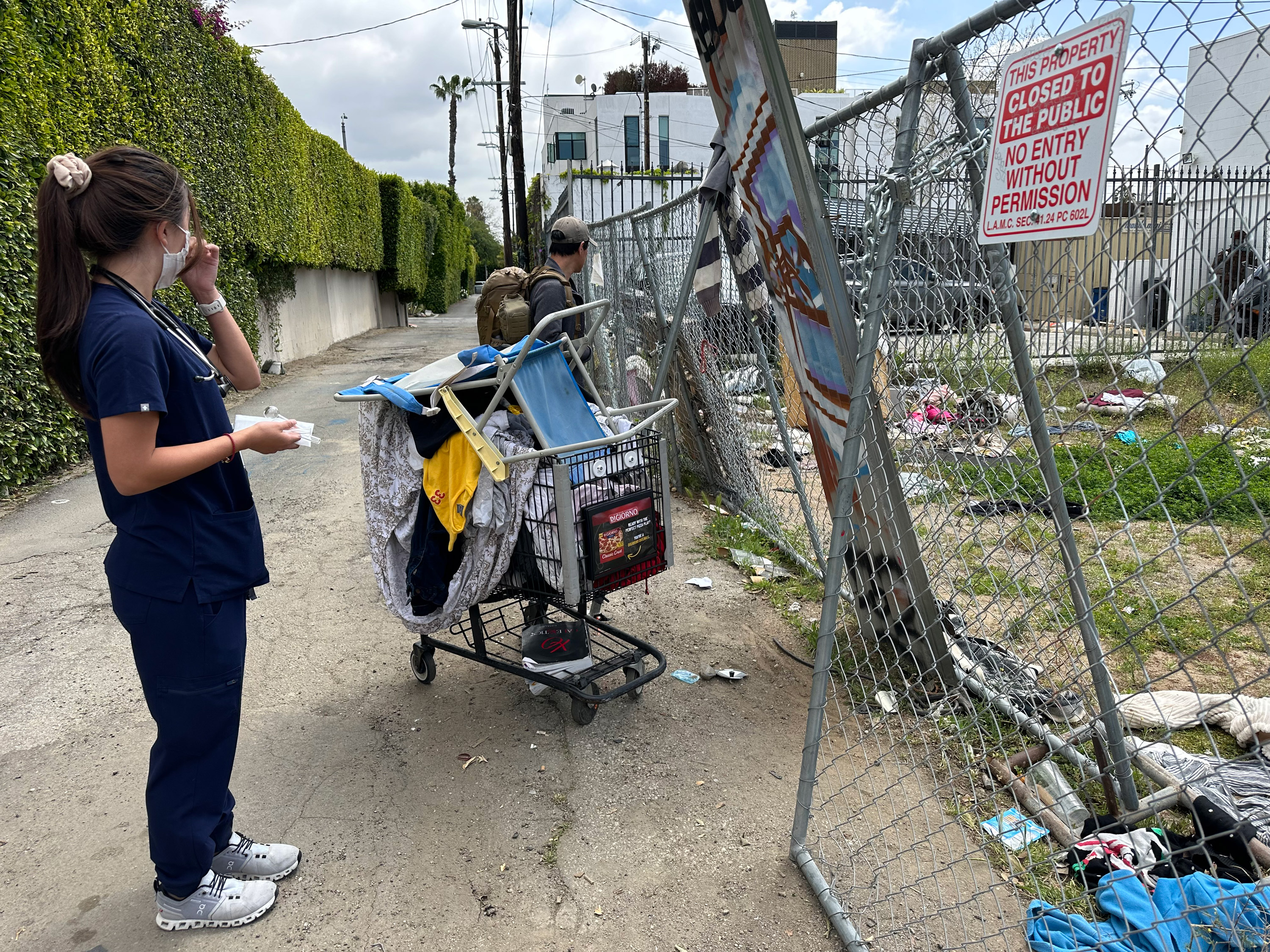 A photo of a medic in scrubs looking down an alleyway. She is next to shopping cart filled with belongings.