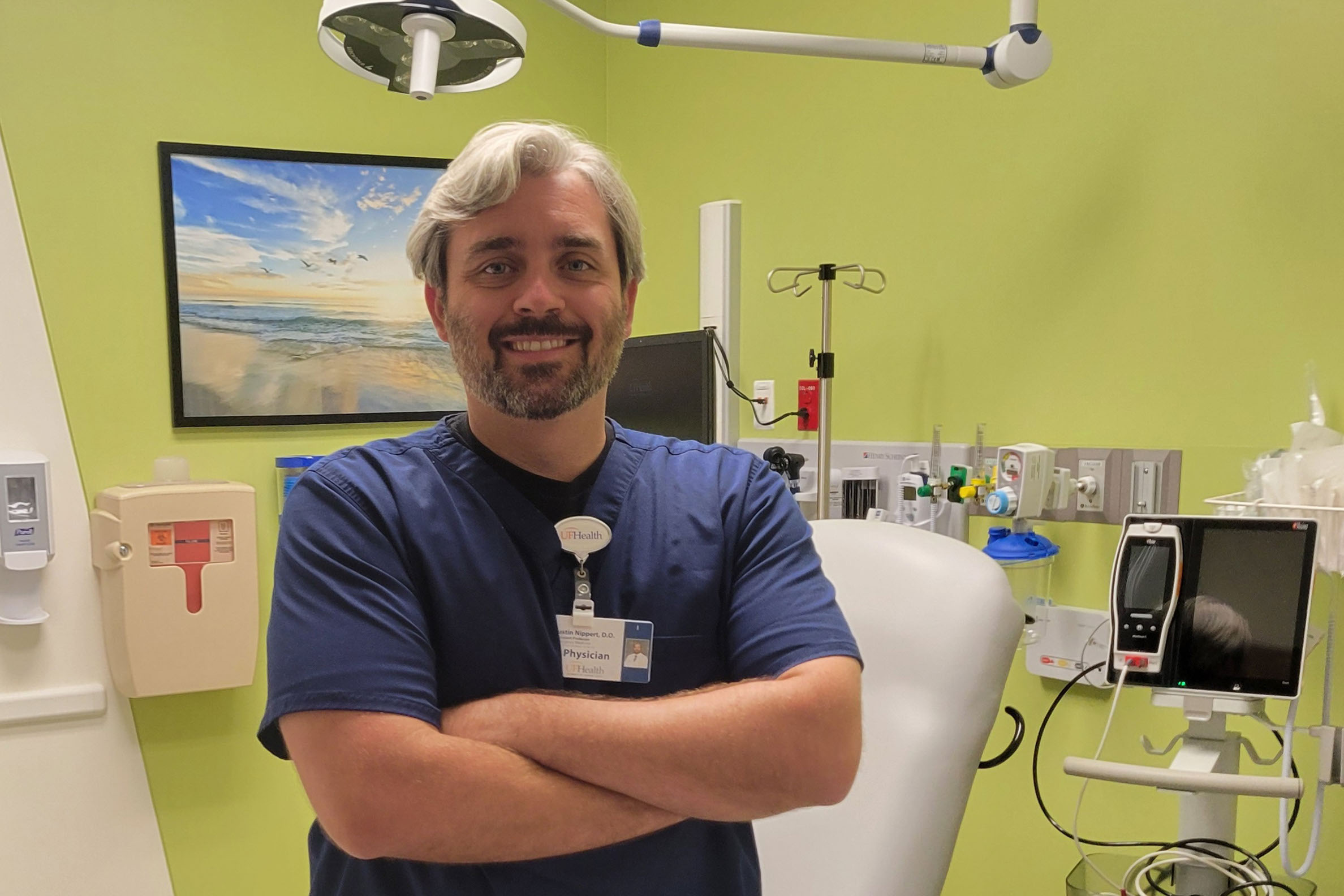 Justin Nippert, an emergency physician, is standing in a medical room. He has his arms crossed and smiles broadly, facing the camera.