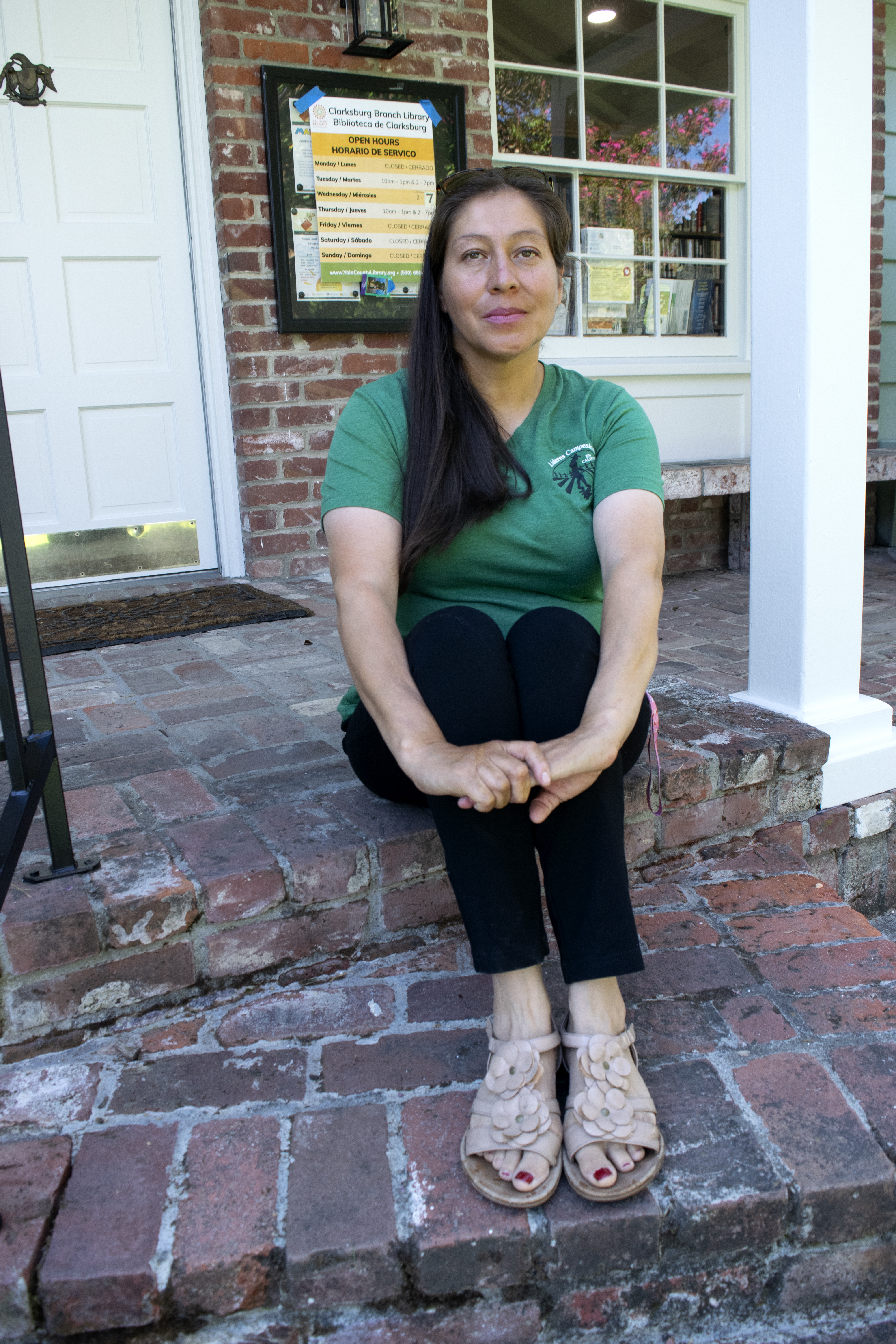 A woman with dark hair and a green t-shirt sits on the floor outside, holding her knees