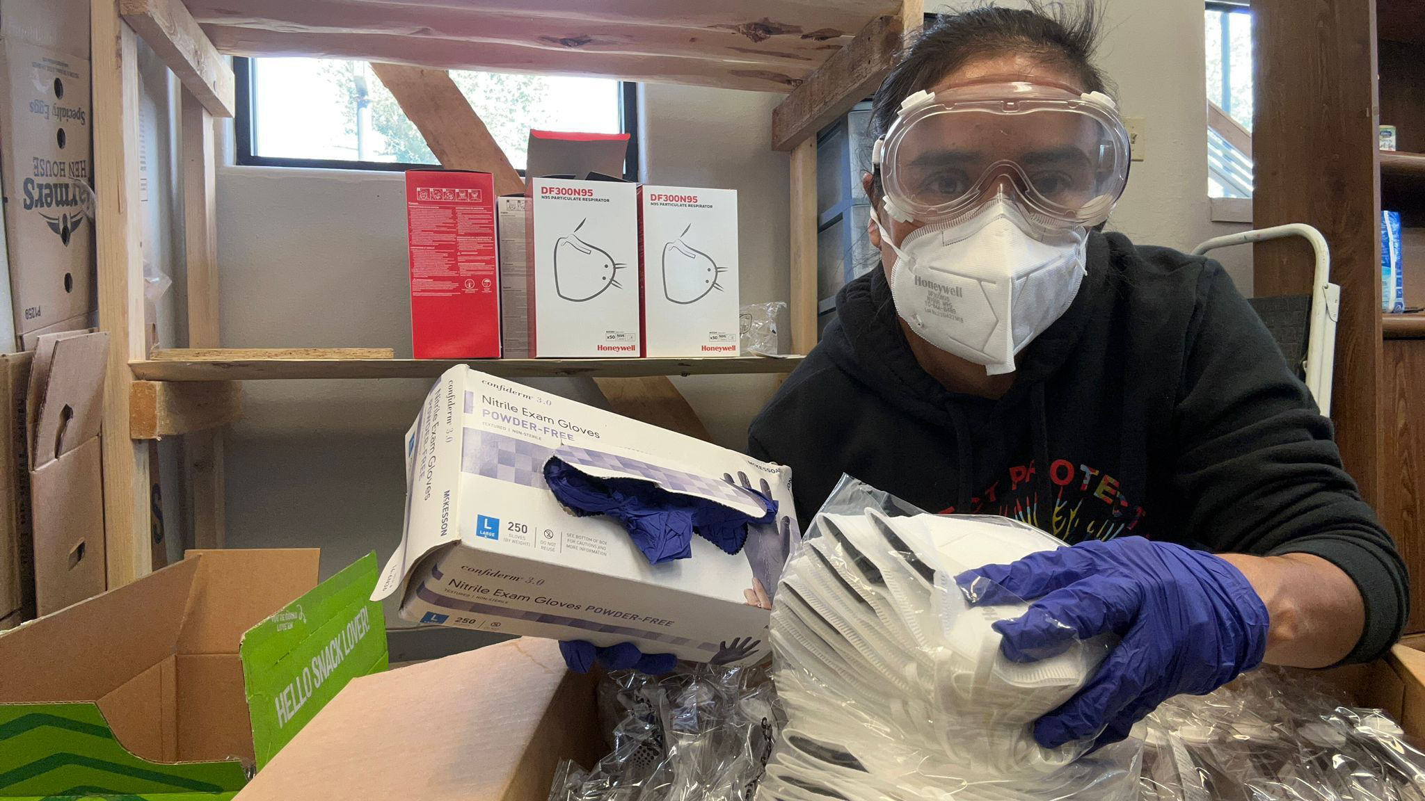 A woman wearing gloves, plastic goggles, and and N95 mask, holds a box of blue nitrile gloves