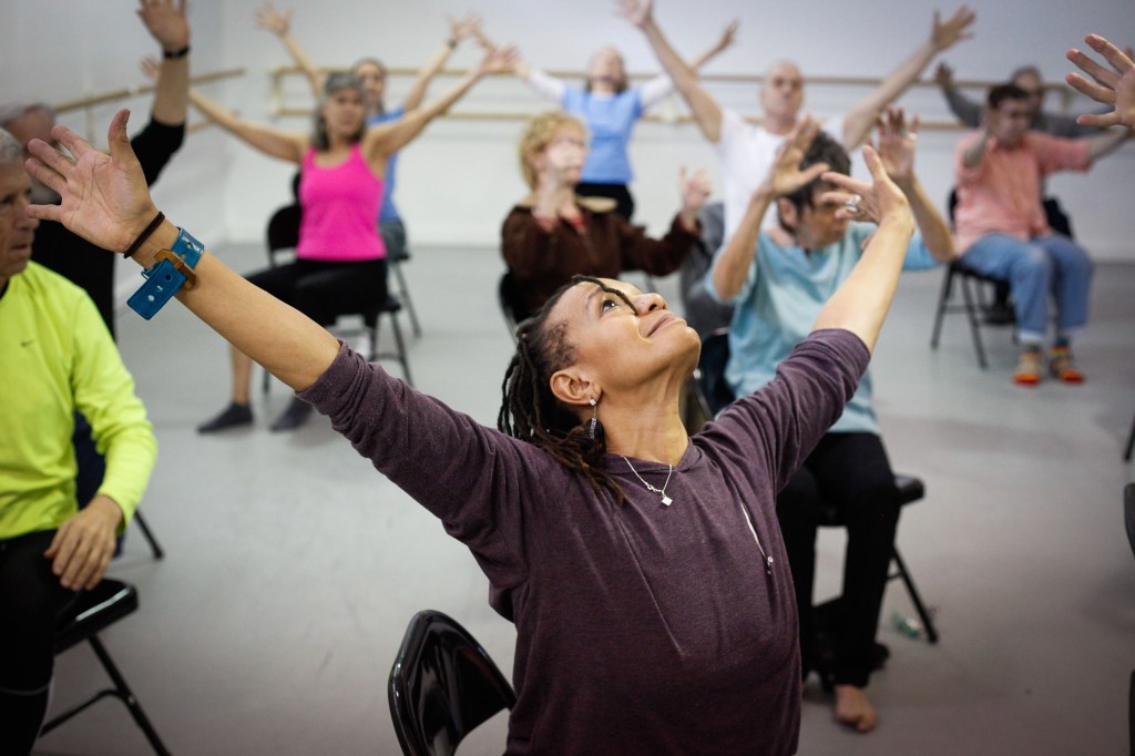 A photograph showing a group of seniors stretching while sitting on foldable chairs.