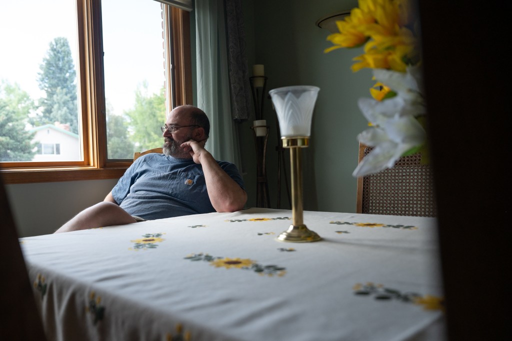 Casey Shively sits for a portrait in his family home. He is sitting at the far end of a table and looks away from the camera, out a window. There are white and yellow lilies on the table, along with a candle holder.