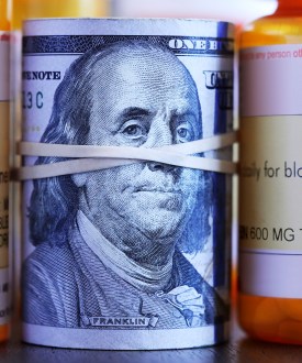 A roll of one hundred dollar bills sits among a row of prescription medication bottles.