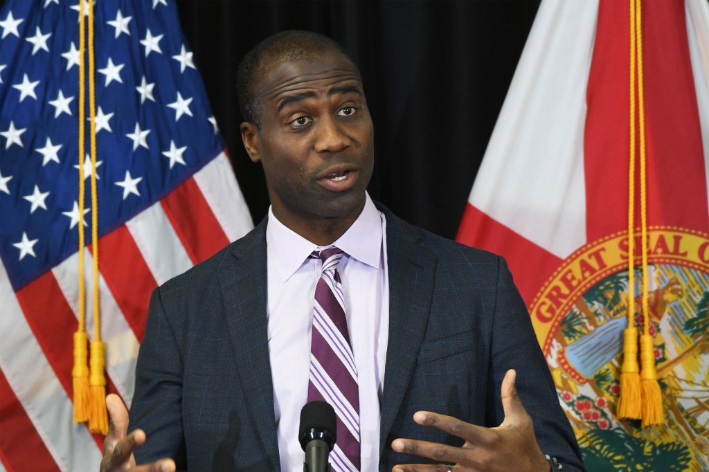 A man in a suit and tie speaks from behind a lectern. A U.S. flag and Florida state flag are behind him.