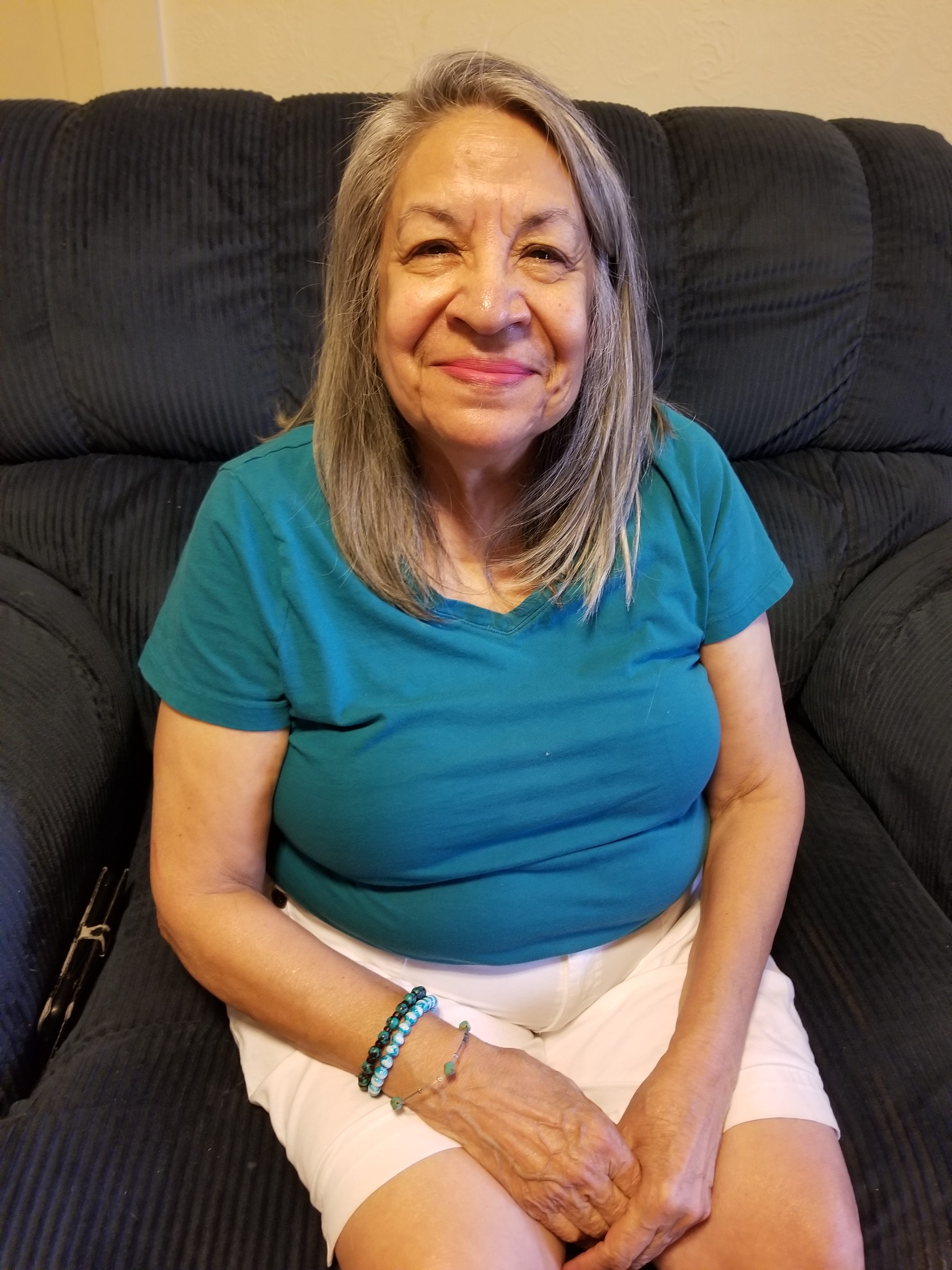 A senior woman in a teal shirt and white shorts sits on a black couch