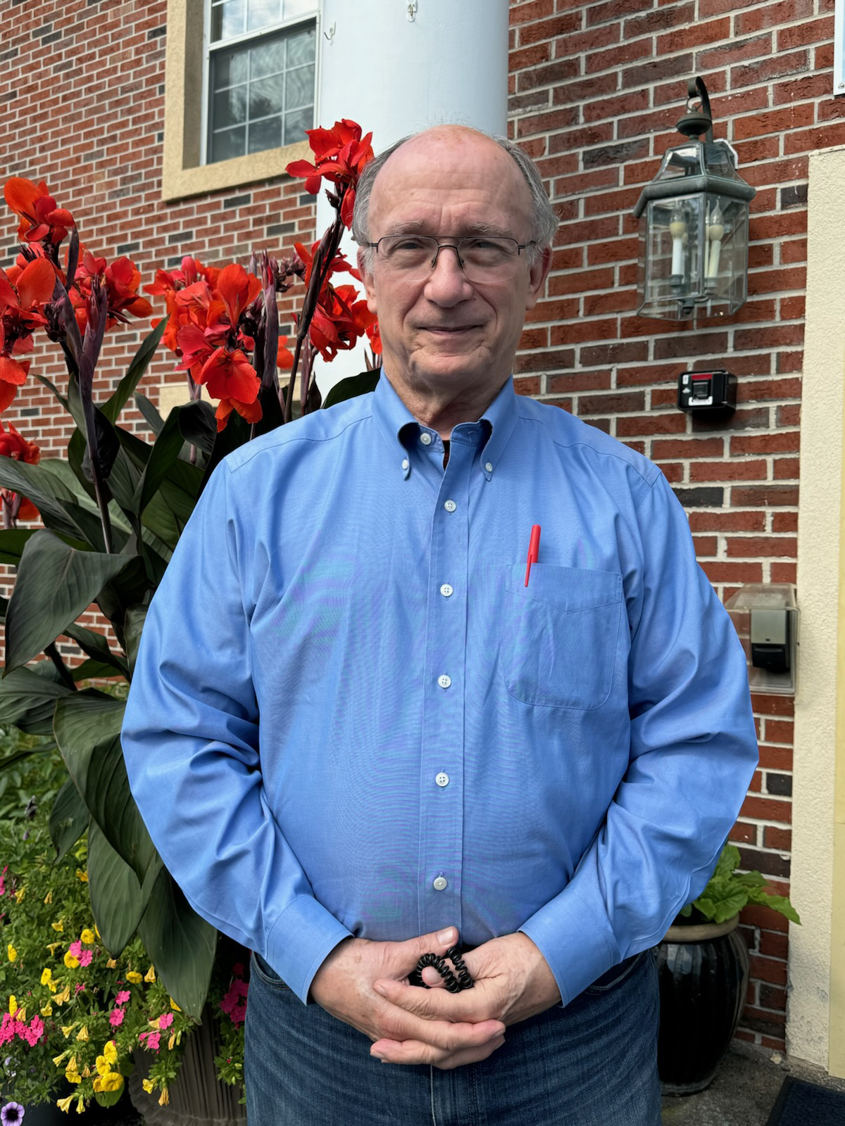 A senior man in a blue button up shirt stands in front of a building a bush with red flowers