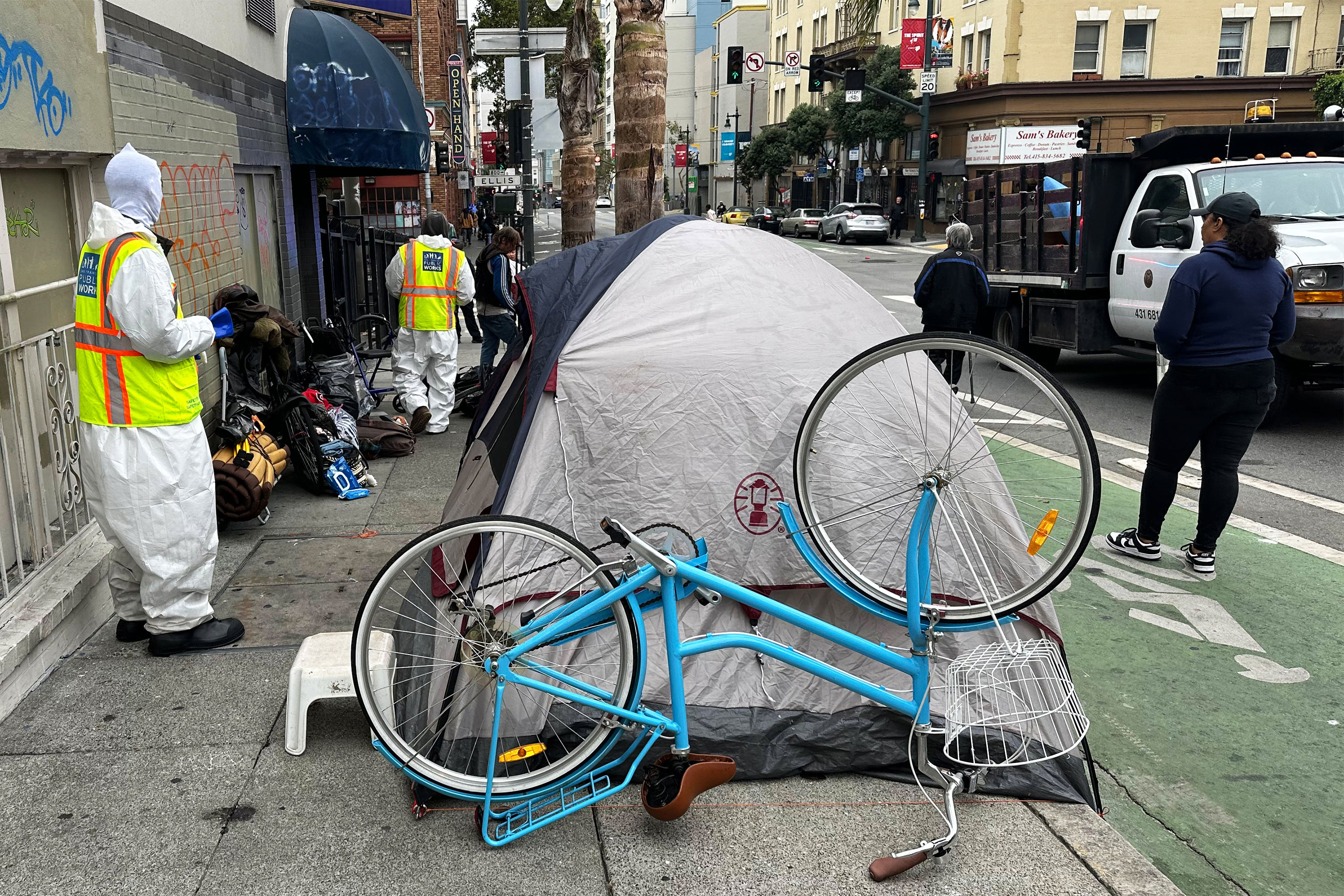 A photo of workers in reflective vests standing by an encampment on the sidewalk.