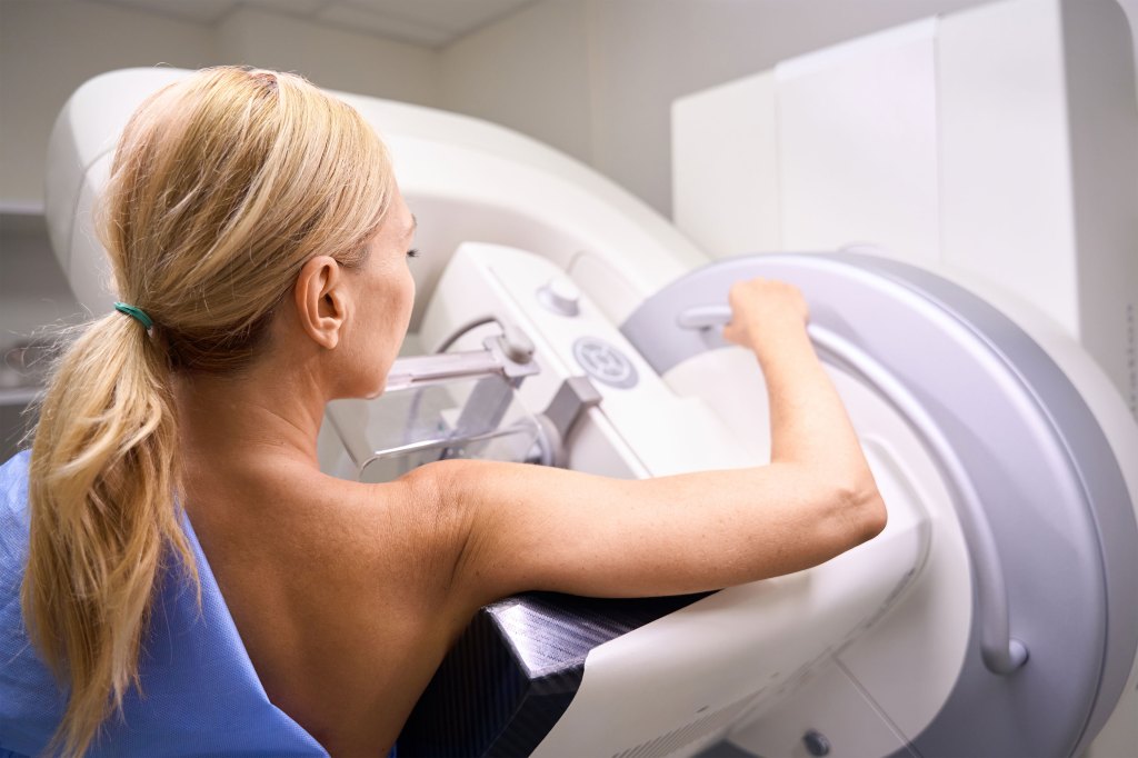 A woman with straight blonde hair stands at a mammogram machine. Her light blue hospital gown is off her right shoulder.