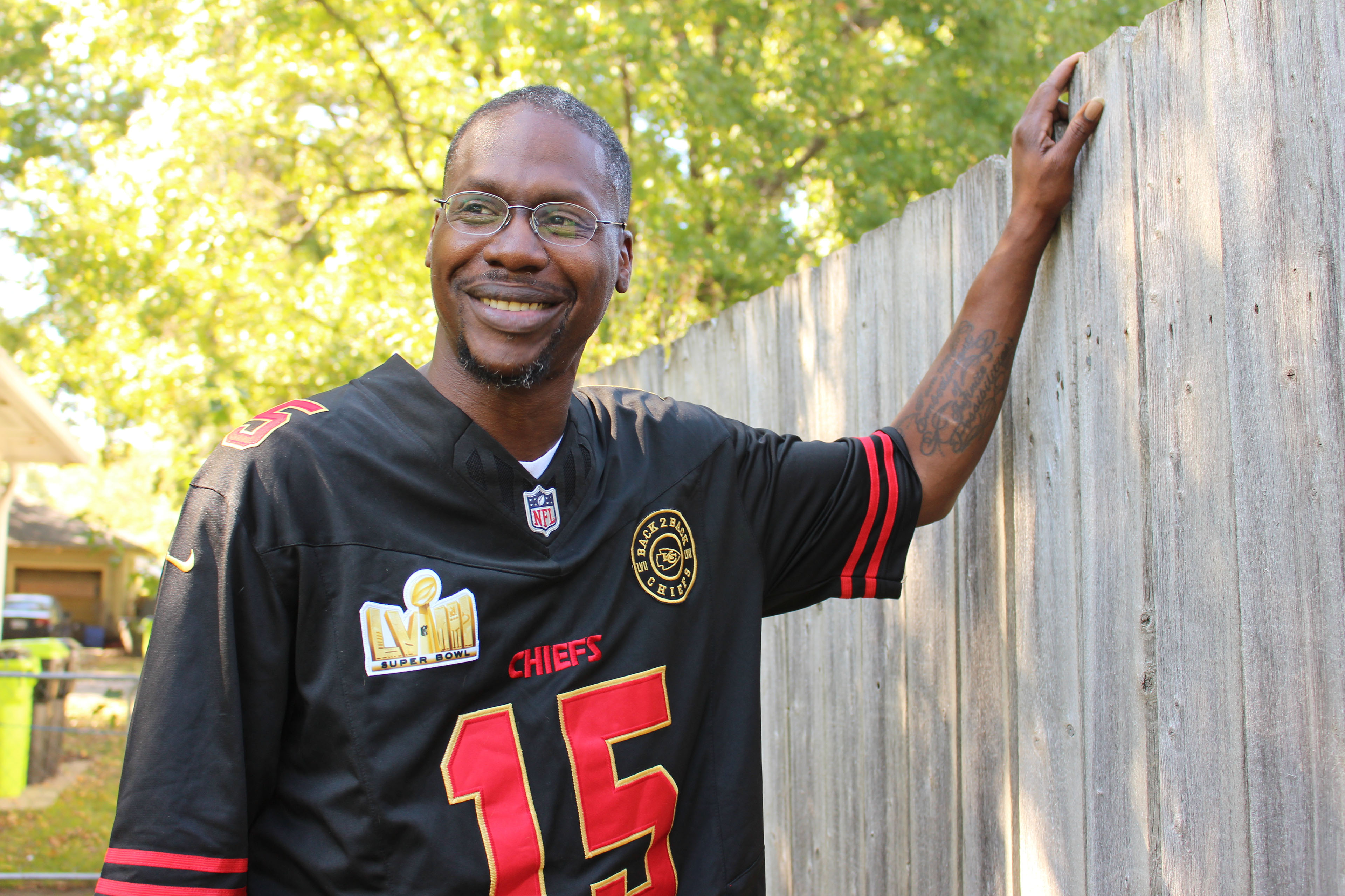 A man wearing glasses and a Kansas City Chiefs football jersey poses for a photo while standing next to a fence