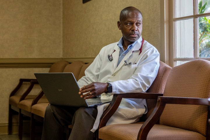 A photo of a Black doctor sitting in his office, looking to the right toward a window.