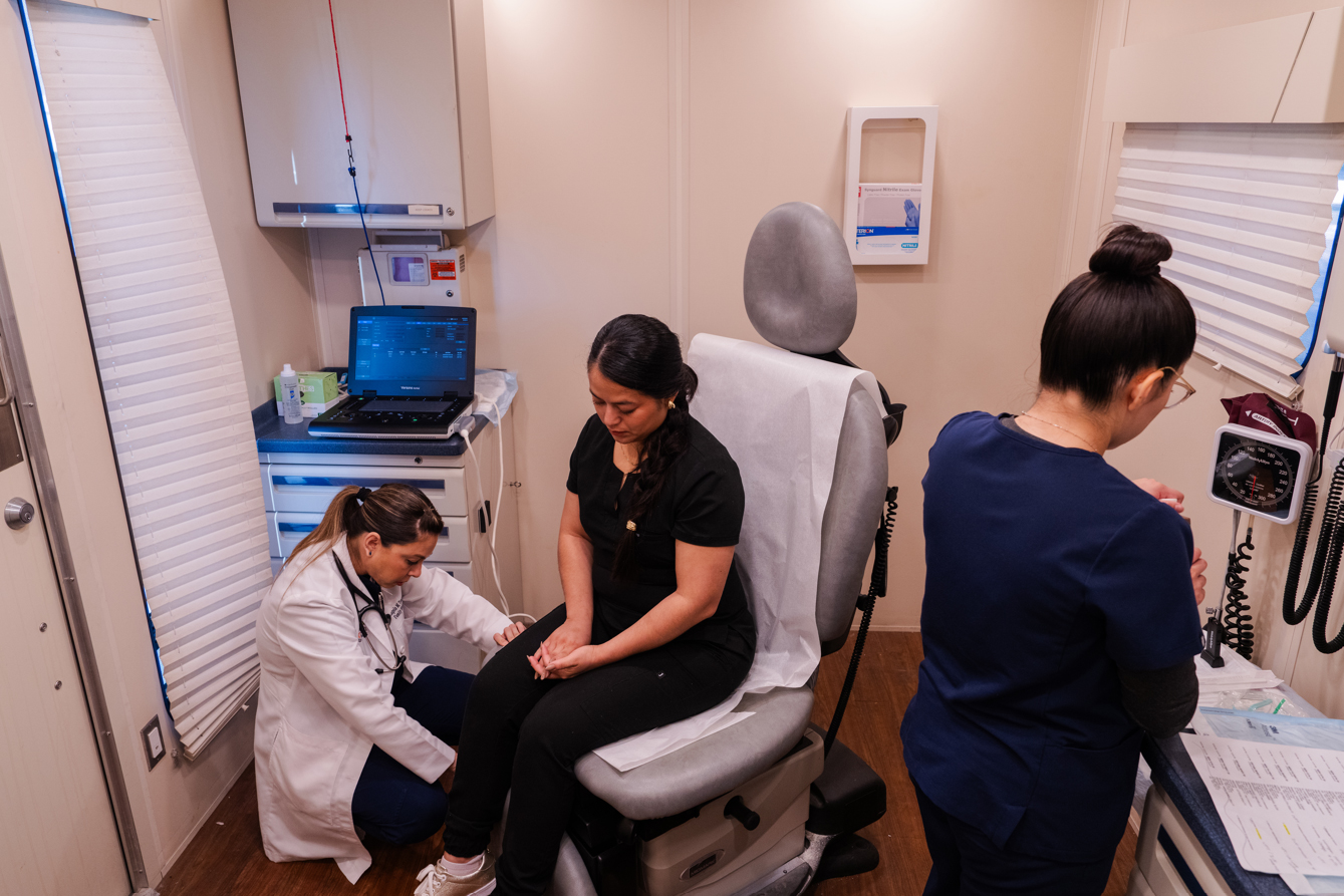 A photo of two medical professionals helping a women sitting in a chair inside a mobile clinic.