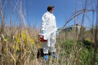 A woman wearing a full-body, white protective suit stands in a field holding an insulated plastic box.