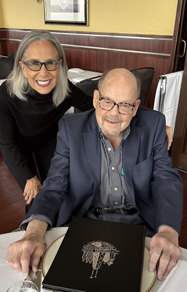 Joy Loverde, an older woman with shoulder length gray hair and glasses, stands beside Gardner Stern, a senior man wearing glasses and a suit jacket. They are sitting down at a table with a white tablecloth.