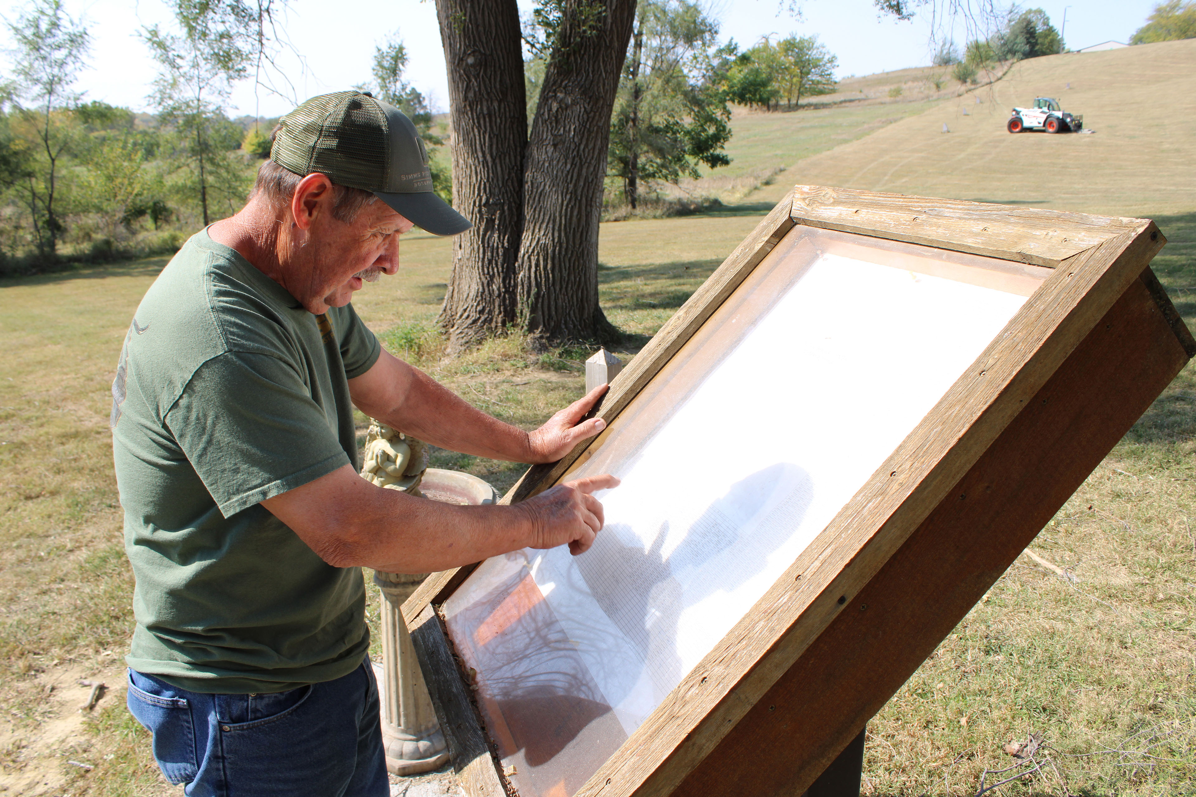 A man is standing beside a wooden sign board and pointing to something on it. He is outdoors on cemetery grounds on a sunny day.