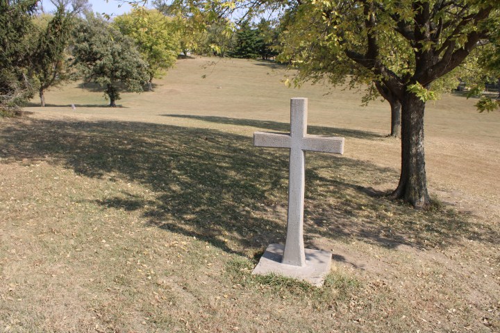 A photograph showing a stone, weathered, 6-foot-tall cross in a graveyard on a sunny day.