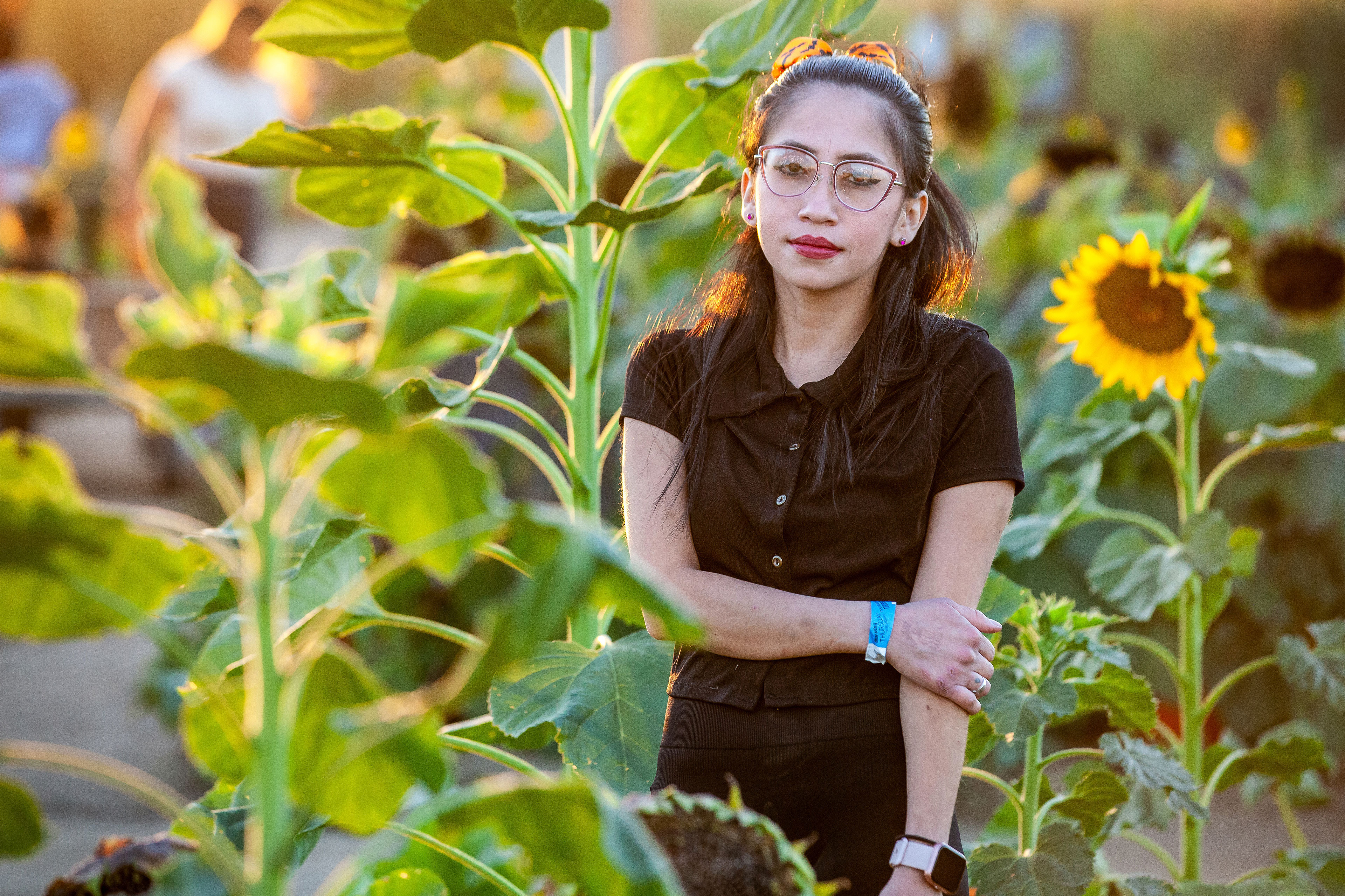 A woman dressed in a black shirt and pants stands in a field of sunflowers and looks at the camera.