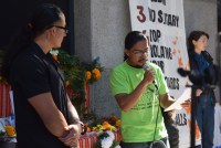 A man in a green shirt and glasses speaks into a microphone in front of an altar of flowers and photos