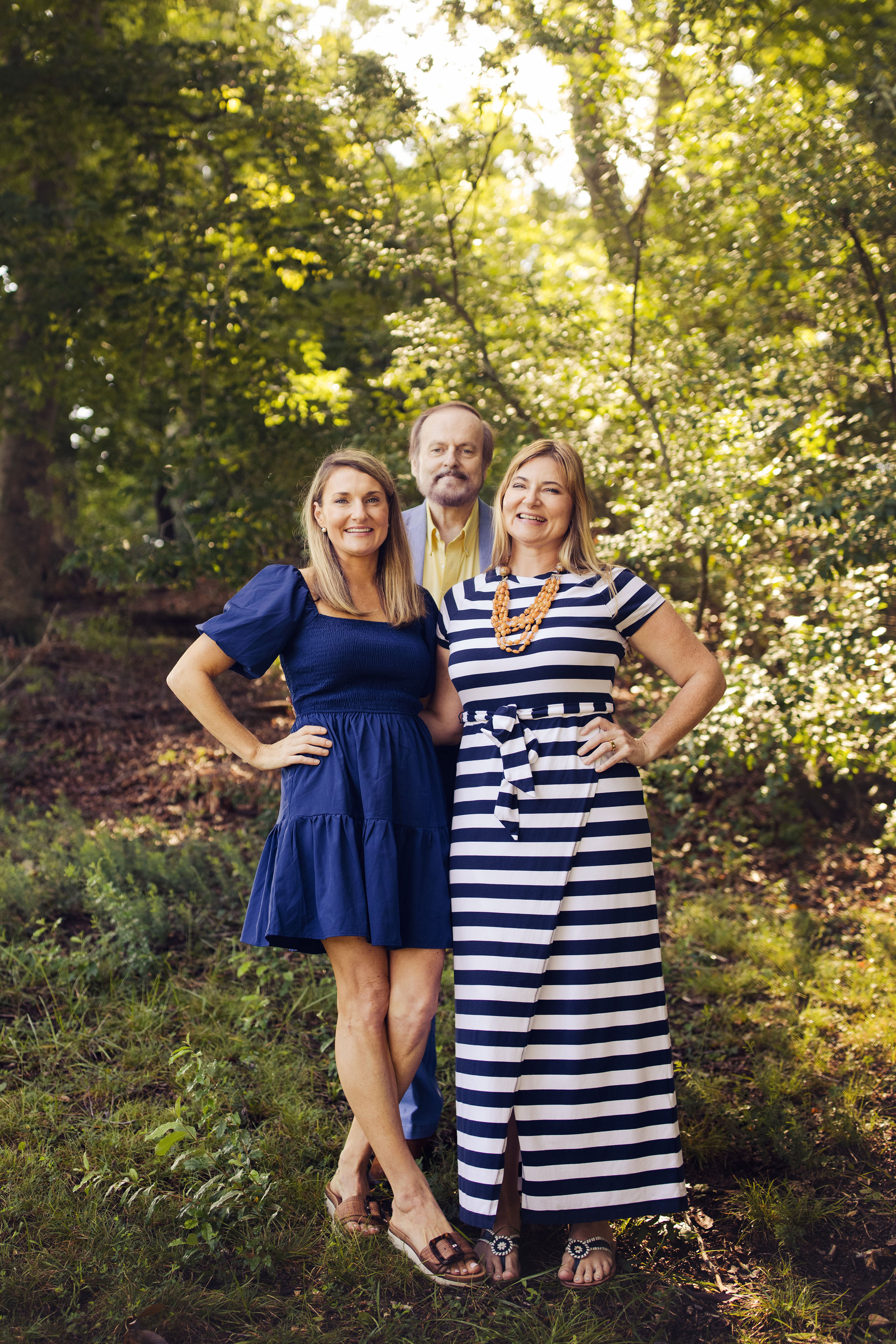 A portrait of a senior father standing with his two adult daughters in a sunlit forest.