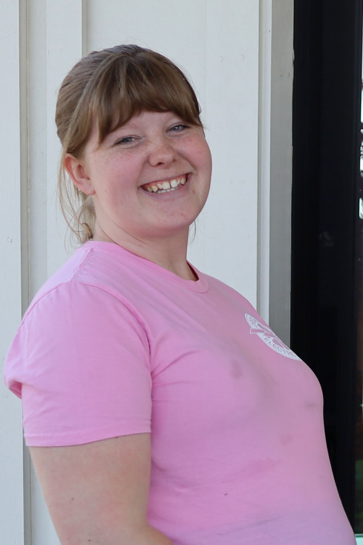 A portrait of a 27-year-old woman wearing a pink shirt. She smiles at the camera.