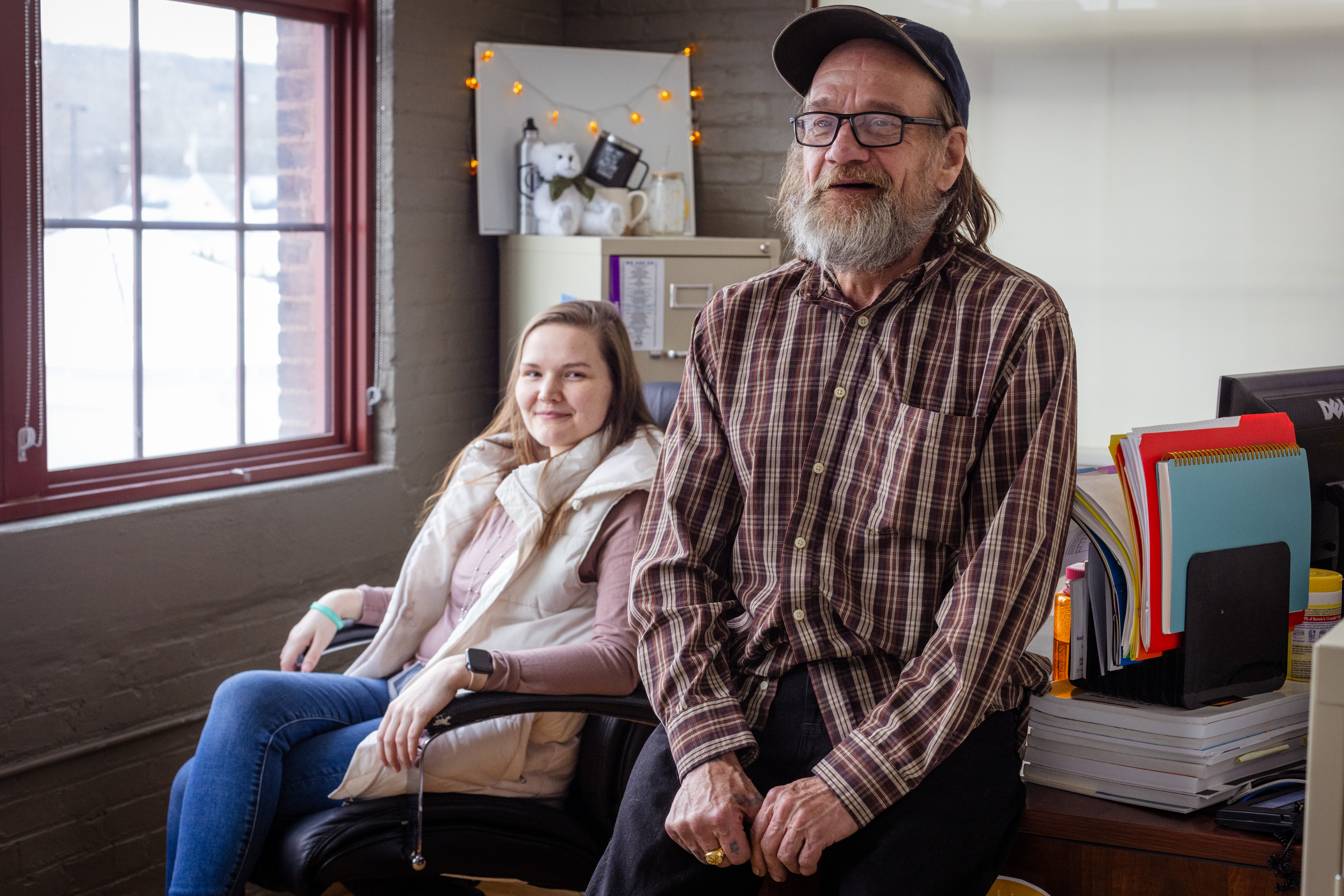 A smiling man with a bushy gray beard stands, leaning against a desk, while a woman sit in a desk chair behind him with her legs crossed.