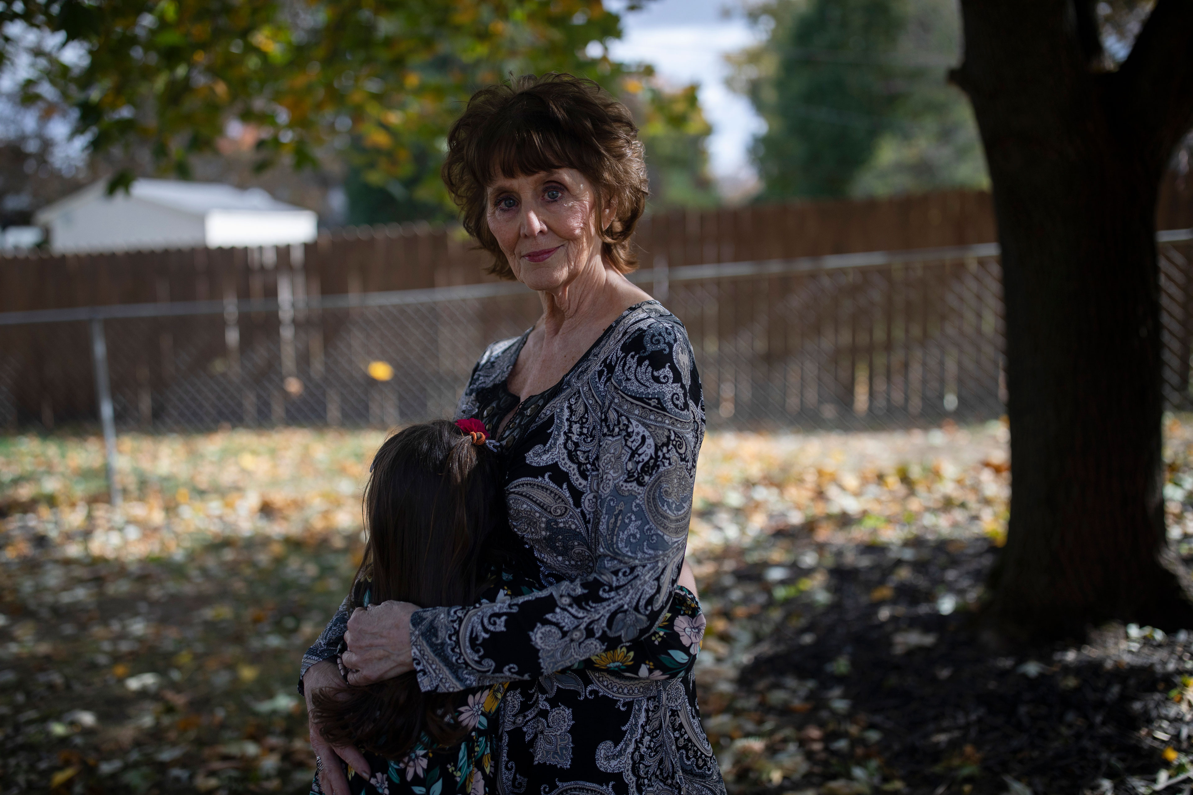 A woman stands outside with her arms around her granddaughter and looks at the camera. The girl's face is turned away from the camera.