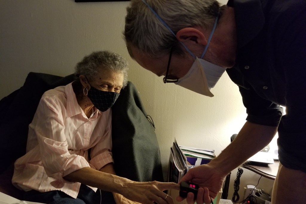 A doctor places a pulse oximeter on a senior woman's index finger. Both are wearing masks.