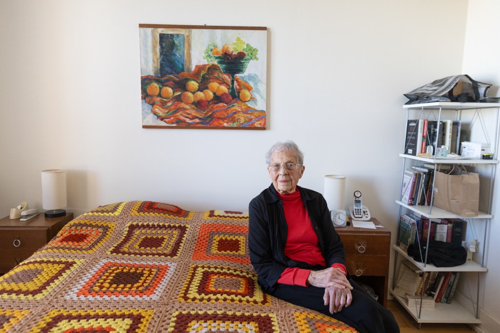 A senior woman wearing a red turtleneck and black cardigan sits on the side of her bed for a portrait. The woven blanket on her bed is made of orange, yellow, and red yarn, which matches the painting of oranges that hangs above her bed.