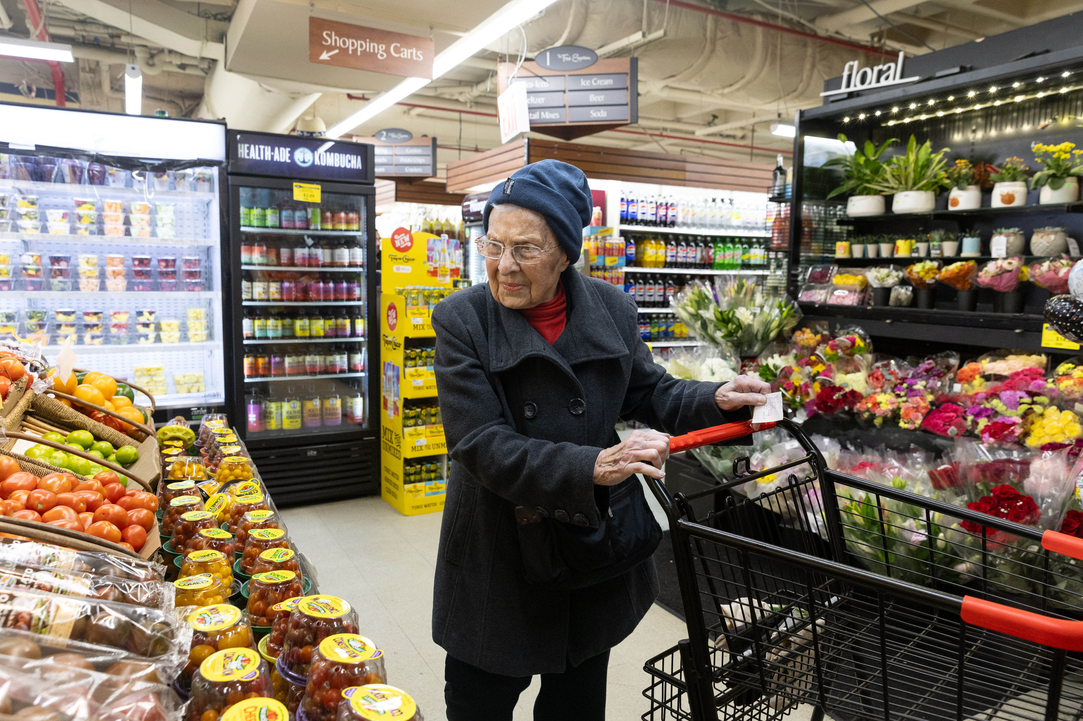 A photo of a senior woman wearing winter clothes while pushing a cart in a grocery store.