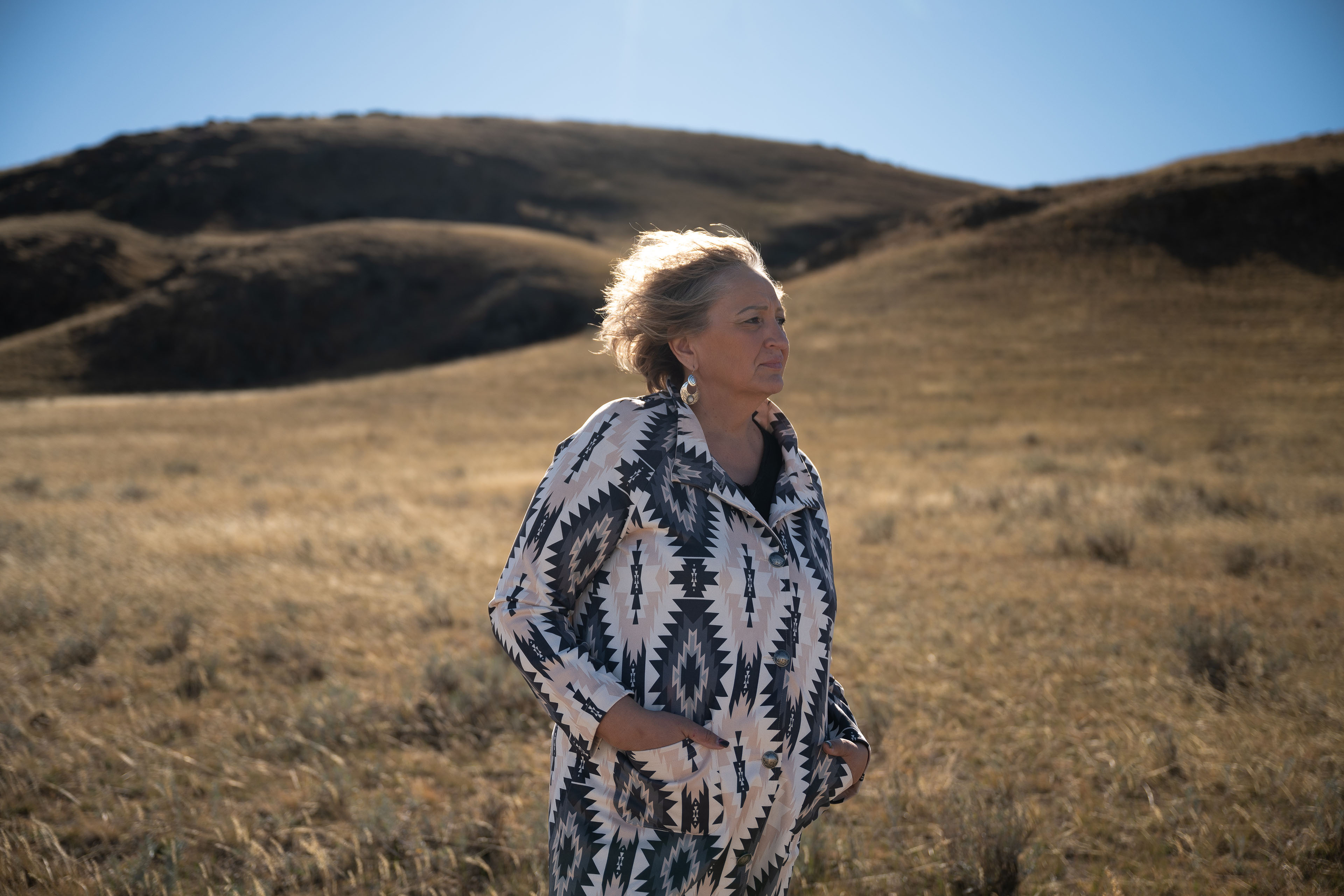 A portrait of a middle-aged woman standing in a grassy field with mountains in the background on a sunny day.