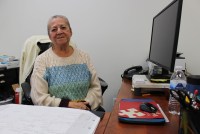 A middle-aged woman sits at a computer desk and smiles in the direction of the camera.