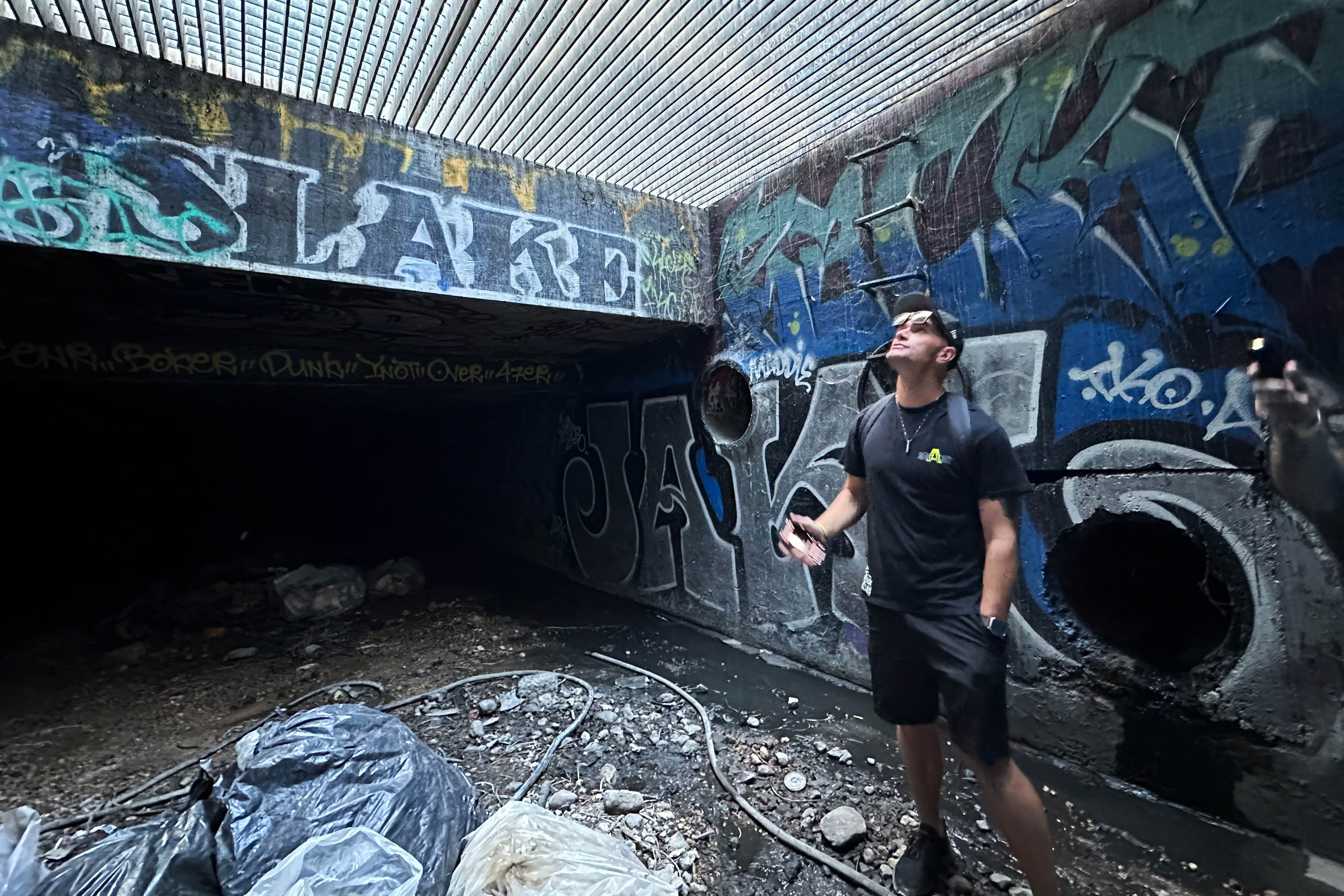 A man, standing in an underground tunnel, looks up at a grate where sunlight filters through.