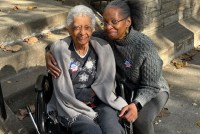 A portrait of two women side-by-side. On the viewer's left is a senior woman in a wheelchair. On the right is a middle-aged woman who has her arm around the senior woman as she kneels beside her. They are both smiling warmly.